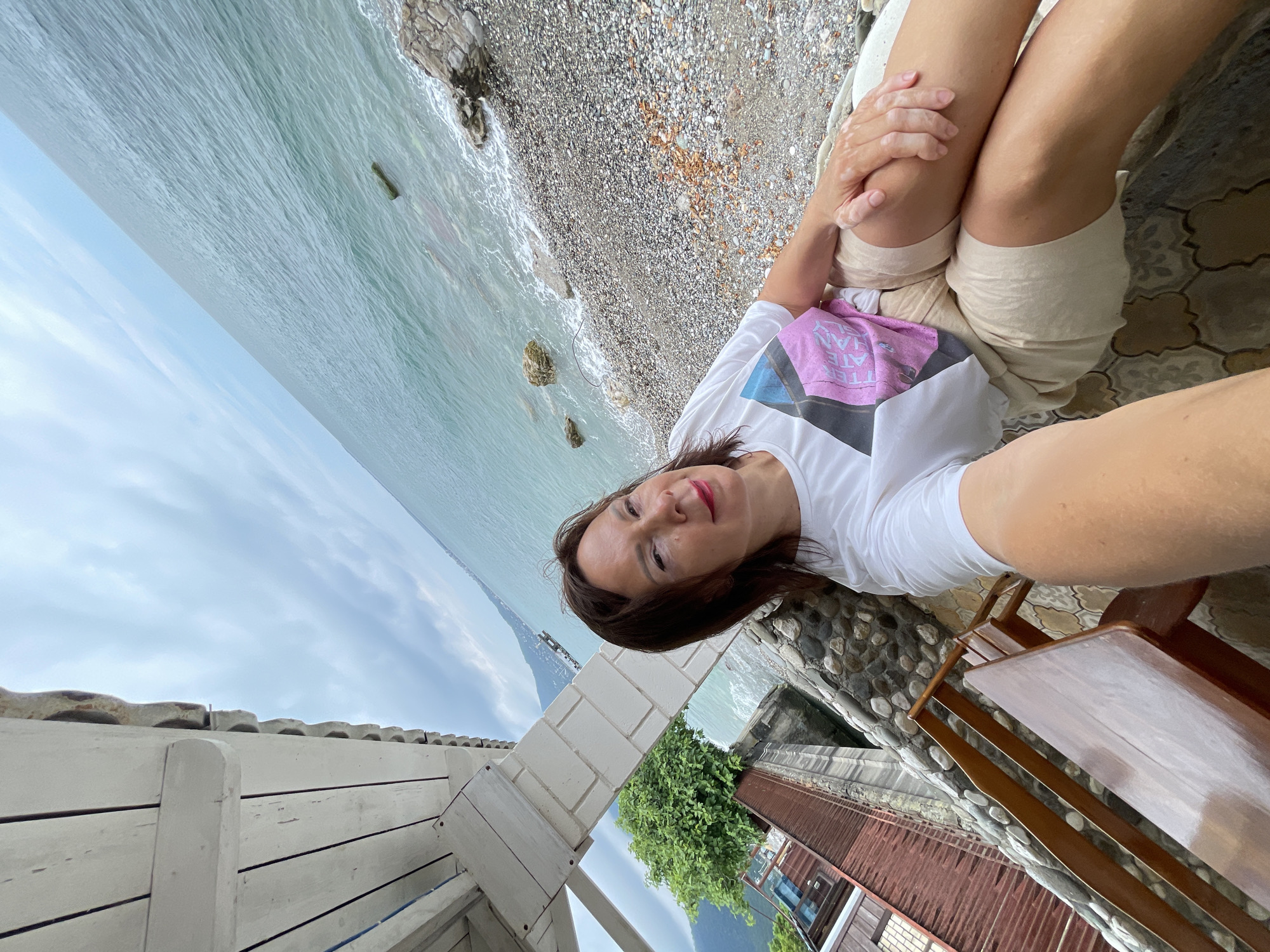 A woman is taking a selfie outdoors, sitting on a bench. She is wearing a white t-shirt with a pink and black graphic and beige shorts. She has brown hair and is wearing red lipstick. The background shows the sea and a pebble beach. To the left of the frame is a wooden structure with a white roof and a stone wall. Further to the left, there is a red-roofed building and a green tree. In the distance, mountains and the coastline are visible. The location is Abaata, Abkhazia.