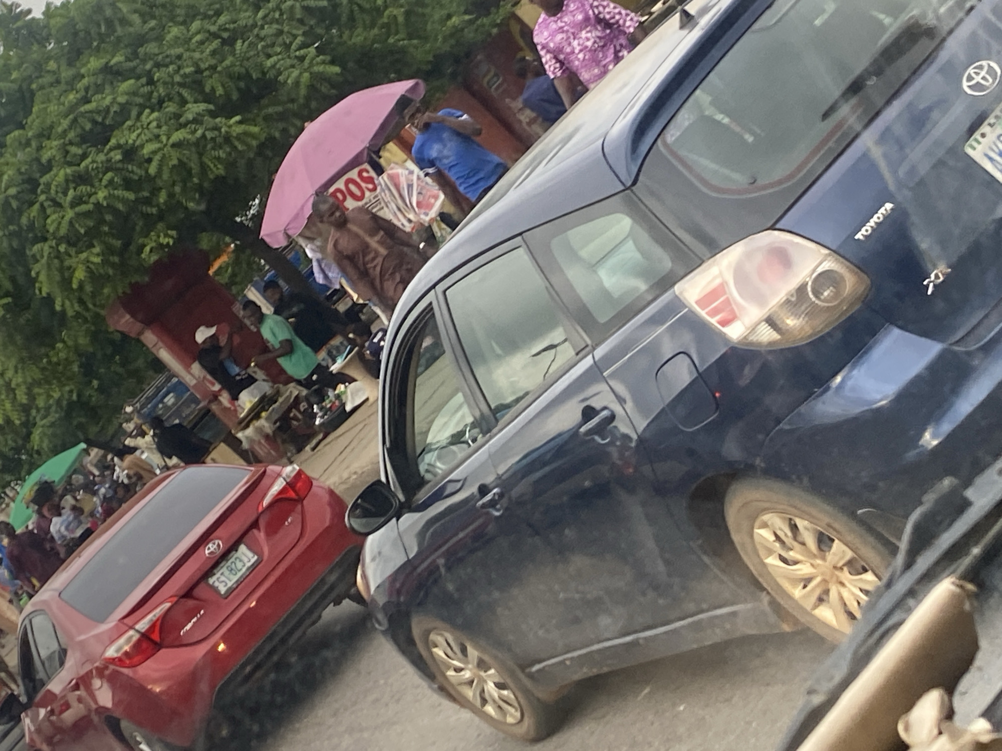 The image depicts a street scene in Lagos, Nigeria, likely during daytime. A red Toyota Corolla car is parked on the left, with its license plate reading "ST 823 JJ". Behind it, a busy market area is visible with several people and stalls. A blue Toyota car is prominently featured in the foreground, showing its rear-left side. Further in the background, various individuals are seen, some under a pink umbrella. The environment appears to be a typical urban setting in Lagos, characterized by traffic, street vendors, and dense human activity. The overall scene suggests a bustling and active street market atmosphere.