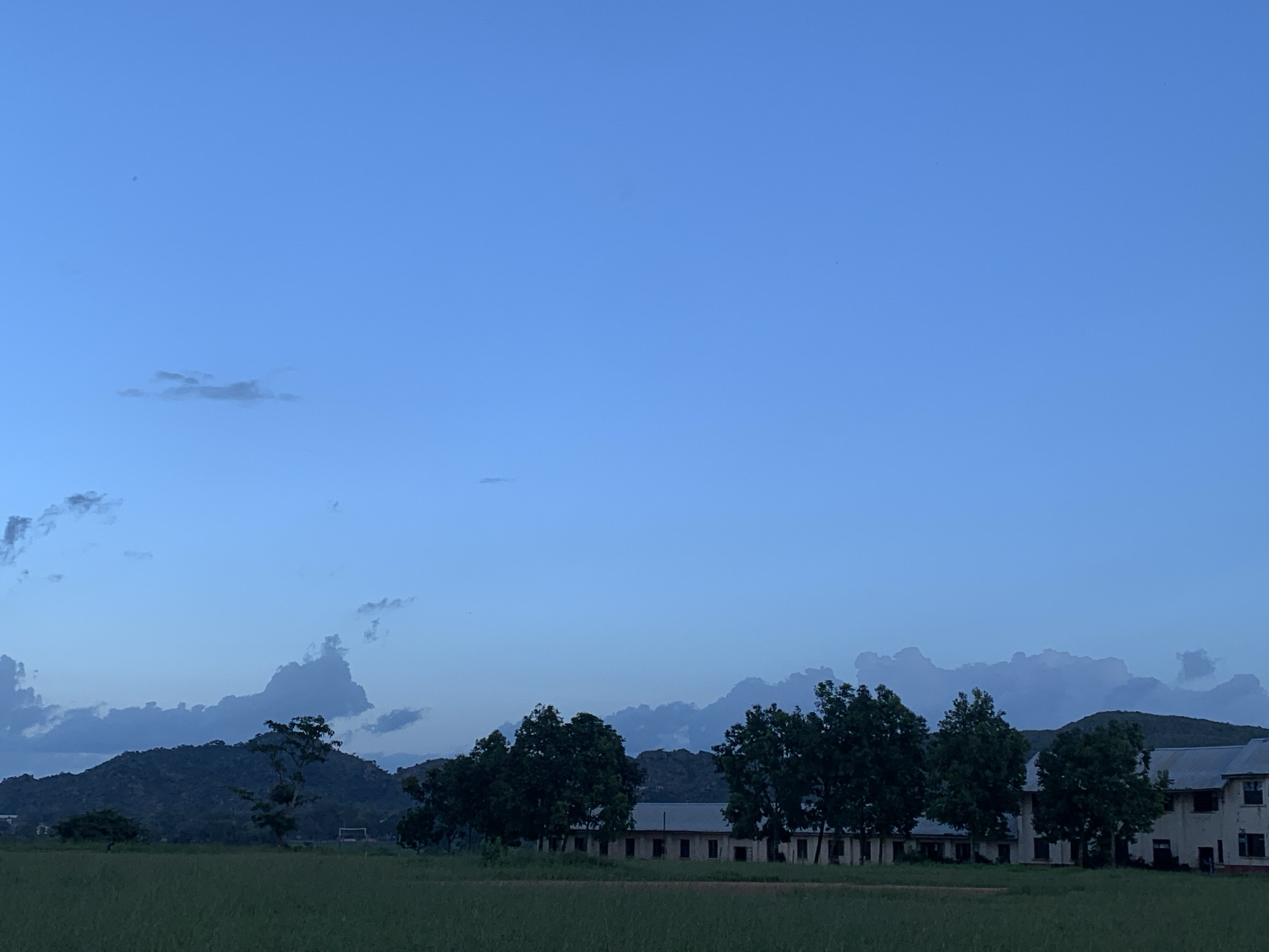 The image depicts a wide, open field in the city of Kuru Karama, Nigeria, under a twilight sky. In the foreground, a lush green field extends across the entire width of the frame. Behind the field, a line of trees stands out, silhouetted against the subdued lighting. Further back, a low-rise building complex with a tiled roof and numerous windows stretches horizontally. In the distance, a range of dark, undulating hills forms the backdrop, with several scattered clouds adding texture to the serene sky. The overall atmosphere is calm and still, characteristic of early morning or late evening light.