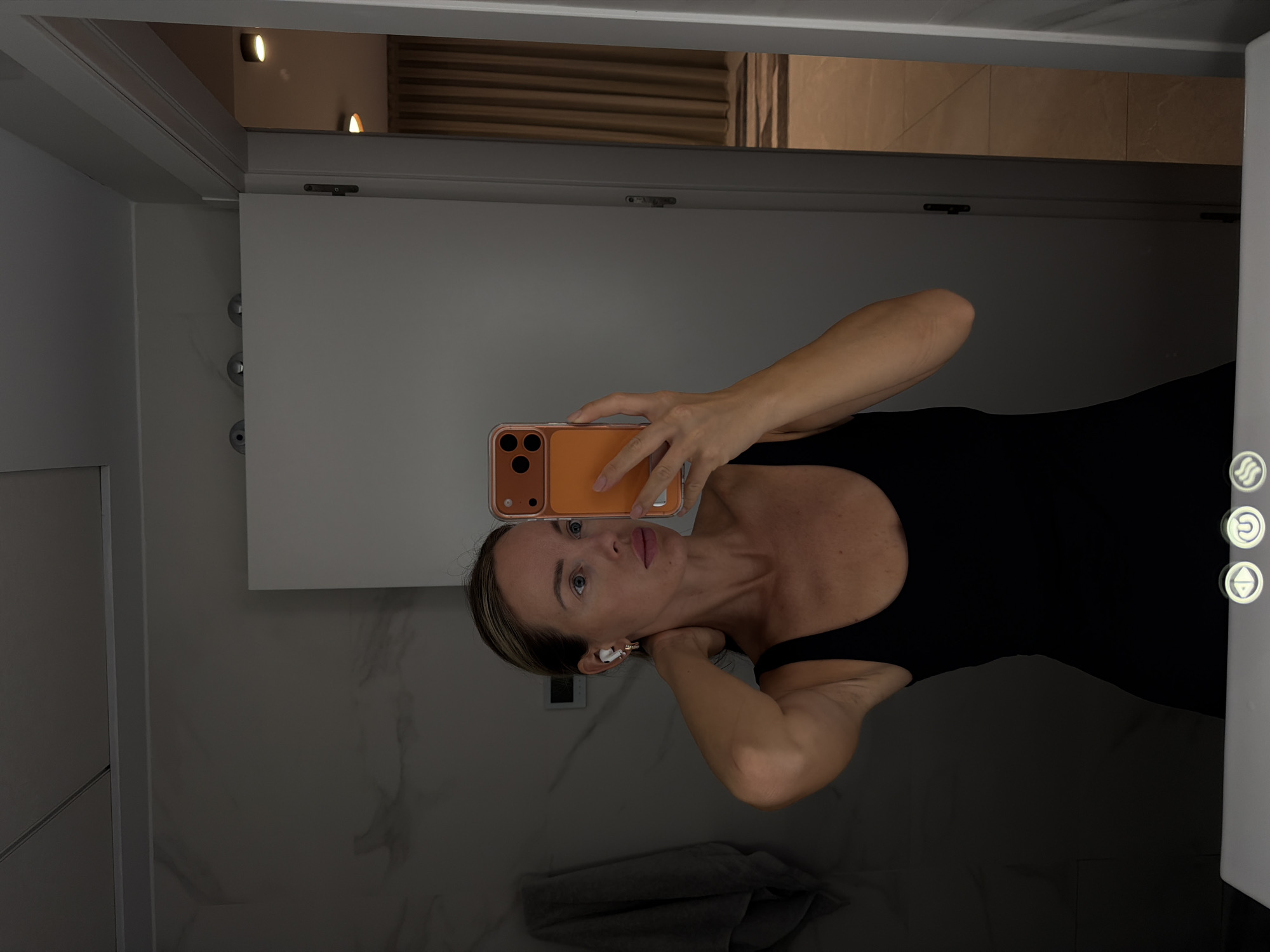 A woman with blonde hair, wearing a black tank top and white wireless earbuds, takes a mirror selfie in a bathroom. She is holding an orange smartphone in her right hand, obscuring her face. Her left hand is touching the back of her neck. The background shows a marble-tiled wall, a closed white door, and a glimpse of a room with beige curtains. The location is the city of Finestrat, Spain.