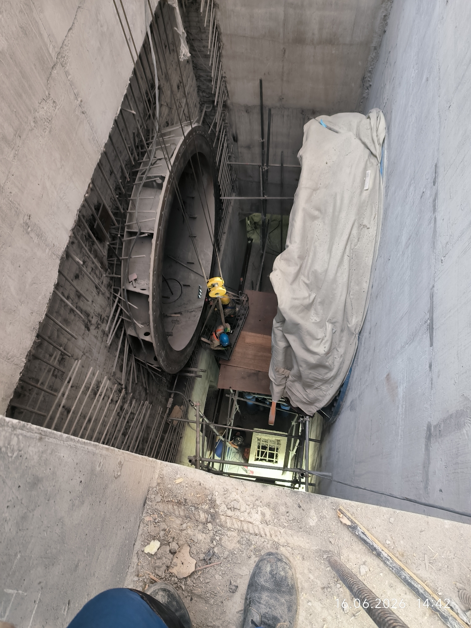The image shows an industrial construction site in an unknown city. A large, circular metal structure, possibly part of a tunnel or large pipe system, is visible in the center of the frame. It is suspended and appears to be undergoing installation or maintenance. Several workers, wearing safety helmets, are present around the structure, some operating machinery, including a yellow hoist. The surrounding environment is characterized by concrete walls, rebar reinforcements, and scaffolding, indicating ongoing construction work. One section of the large metal structure is covered with a light-colored tarp. In the foreground, the feet of a person wearing work boots and blue pants are visible, suggesting the image was taken from a slightly elevated perspective. The overall scene conveys a busy and active construction environment.