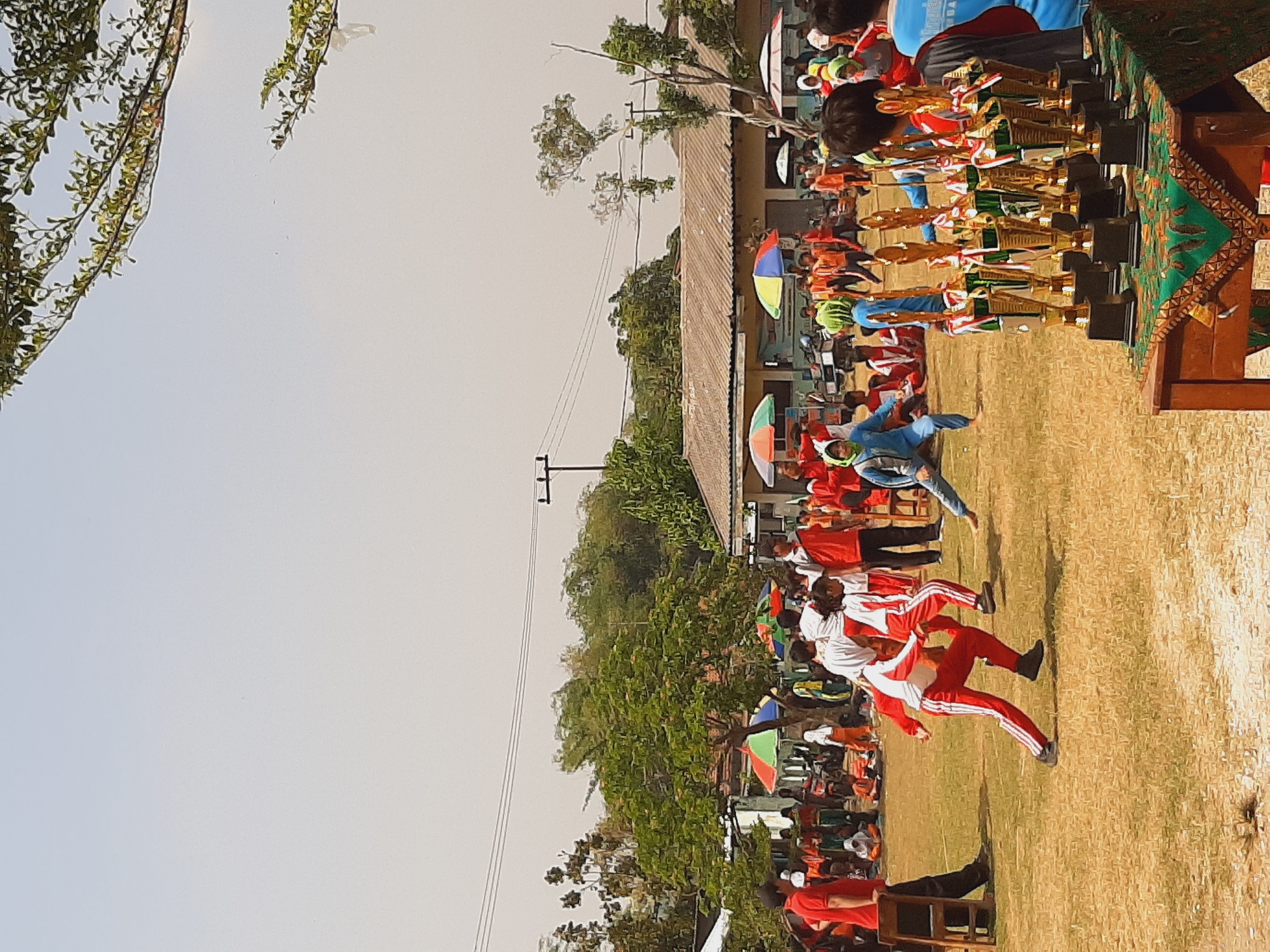 A wide shot captures a daytime outdoor event, likely a sporting competition, taking place on a dry grass field. In the foreground, a collection of trophies sits on a wooden table draped with a patterned green cloth. Beyond the table, several children in red and white athletic wear are participating in an activity, with one in mid-stride. Other participants in similar attire are scattered across the field, some engaged in activities and others observing. Spectators are visible in the background, seated or standing under colorful umbrellas. A building with a corrugated roof and open windows, along with trees and utility poles, forms the backdrop. The sky is a hazy light blue, suggesting a warm and possibly humid climate. The overall scene depicts a community gathering and celebration of athletic achievement in what appears to be a school or local park setting. The image location is identified as the city of Sumberjo, Indonesia.