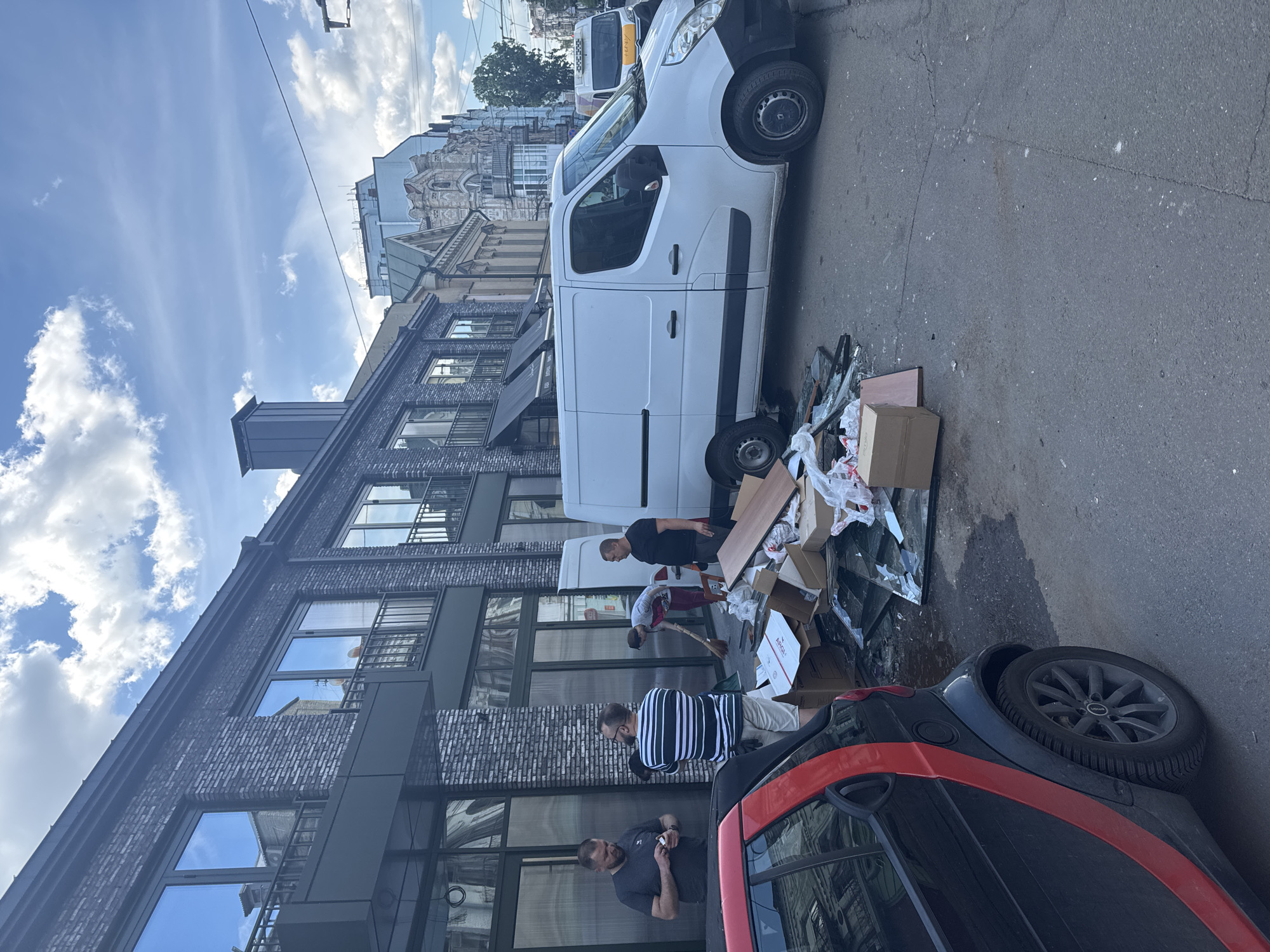 The image shows a street scene in Kyiv, Ukraine. A white van is parked on the side of the street next to a black and red Smart car. Debris, including cardboard boxes, broken glass, and wooden planks, is scattered on the ground near the van. Four men are visible. One man, wearing a black shirt, stands near the debris. Another man in a gray shirt is sweeping the debris. A third man wearing a striped shirt stands near the debris, while a fourth man in a gray shirt appears to be smoking a cigarette and looking at his watch. The buildings lining the street are a mix of modern and older architectural styles. The sky is partially cloudy.