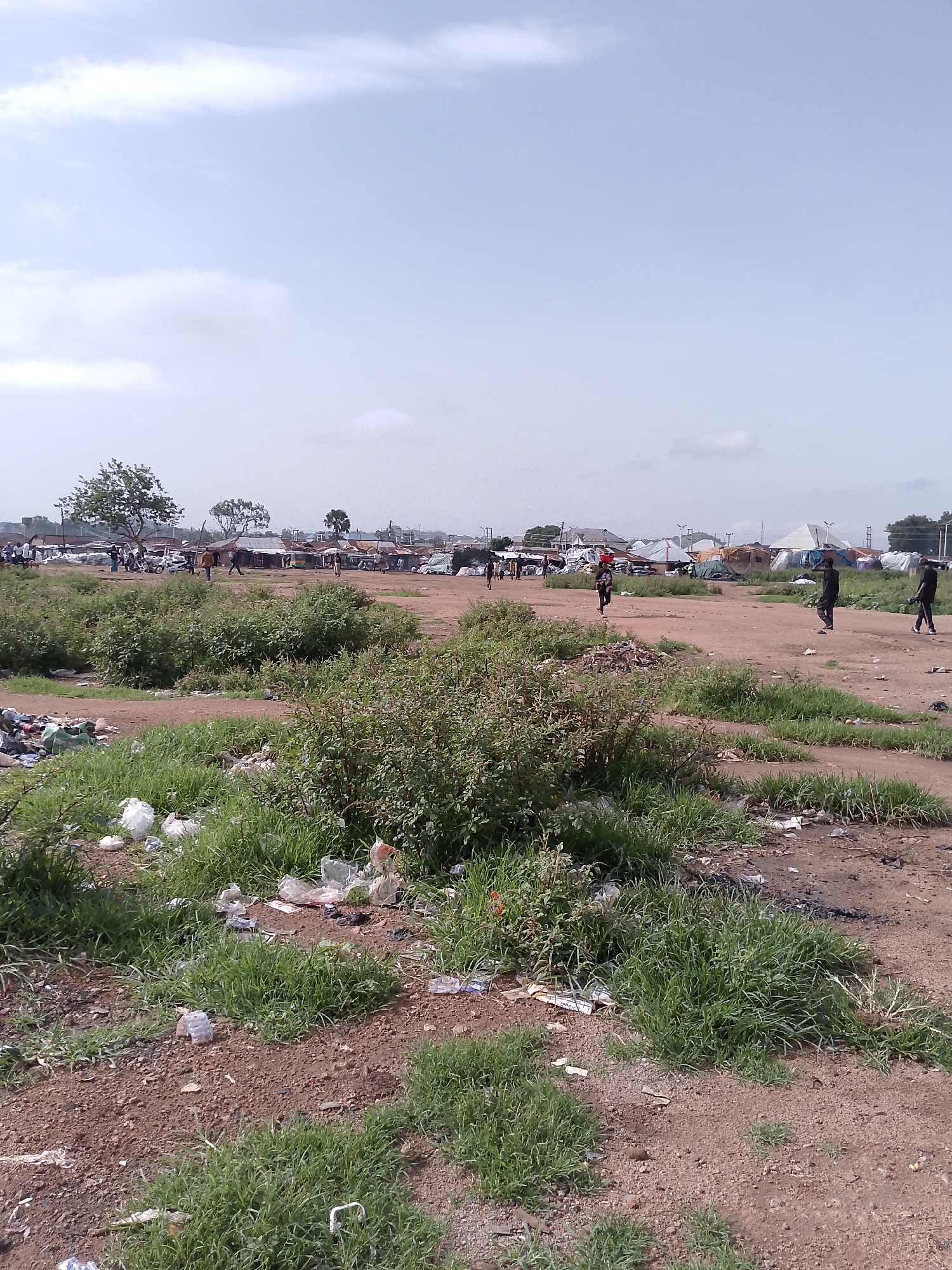 The image depicts an outdoor scene in Bukuru, Nigeria, characterized by a dusty, open area with scattered vegetation and debris. In the foreground, overgrown grass and dry earth are visible, interspersed with plastic waste and other refuse. A cluster of bushes dominates the lower left of the frame.
In the midground and background, the environment appears to be a bustling informal settlement or market. Several makeshift structures, including tents and shacks with corrugated metal roofs, are set up. There are also larger, white tent-like structures in the distance. Various people are walking around, some appearing to be engaged in activity within the settlement. Several motorcycles and a car are parked in the area. The overall impression is one of a lively, if somewhat unkempt, community space. The sky above is overcast with scattered clouds.