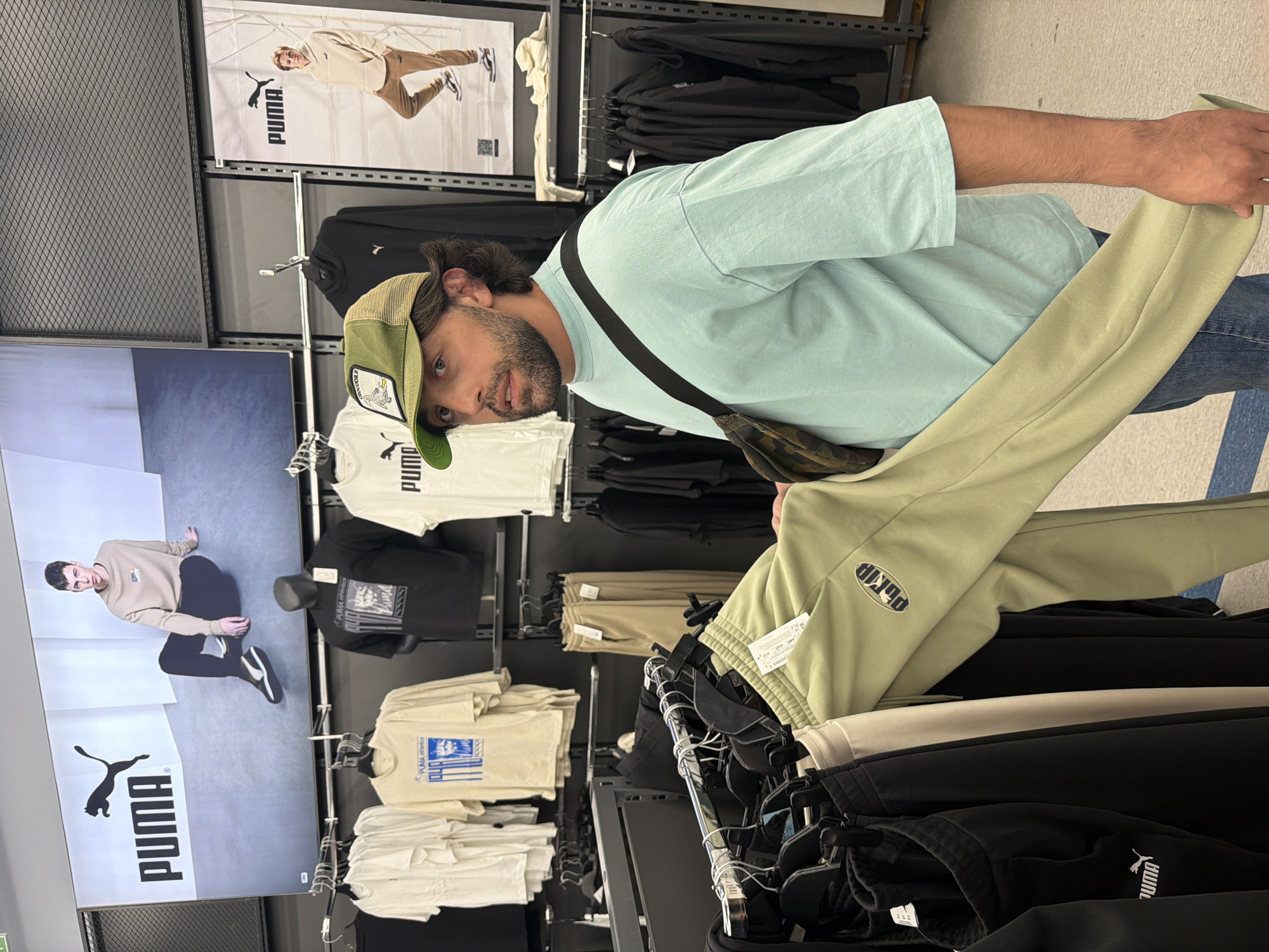 A man stands in a Puma store in Krasnoyarsk, Russia, holding a pair of light green Puma sweatpants. He is wearing a light blue t-shirt, jeans, a green trucker hat, and a crossbody bag. Racks of Puma clothing are visible in the background. Puma advertisements and mannequins are displayed on the walls. The scene is lit with fluorescent lighting.