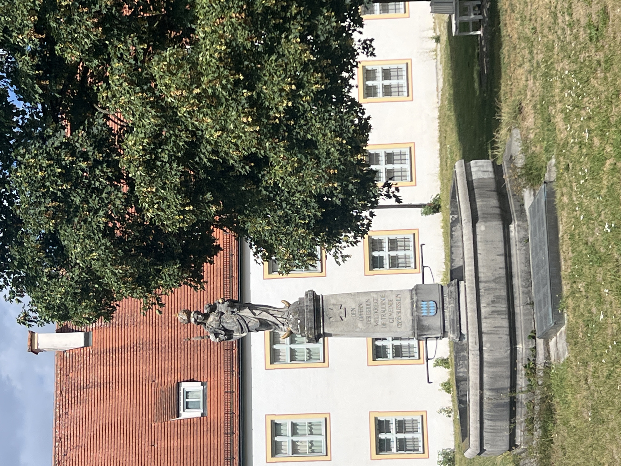 The image shows a stone fountain in front of a white building with a red tile roof. The fountain has a statue of a woman holding a child on top of a pedestal. The pedestal has an inscription that reads "DEN OPFERN DER BEIDEN WELTKRIEGE DIE TRAUERNDE GEMEINDE OBERSCHLEISSHEIM". To the right of the fountain, a large tree with green leaves casts a shadow on the grass. In the background, a white bench is visible. The scene appears to be set in a park or courtyard in Oberschleißheim, Germany.