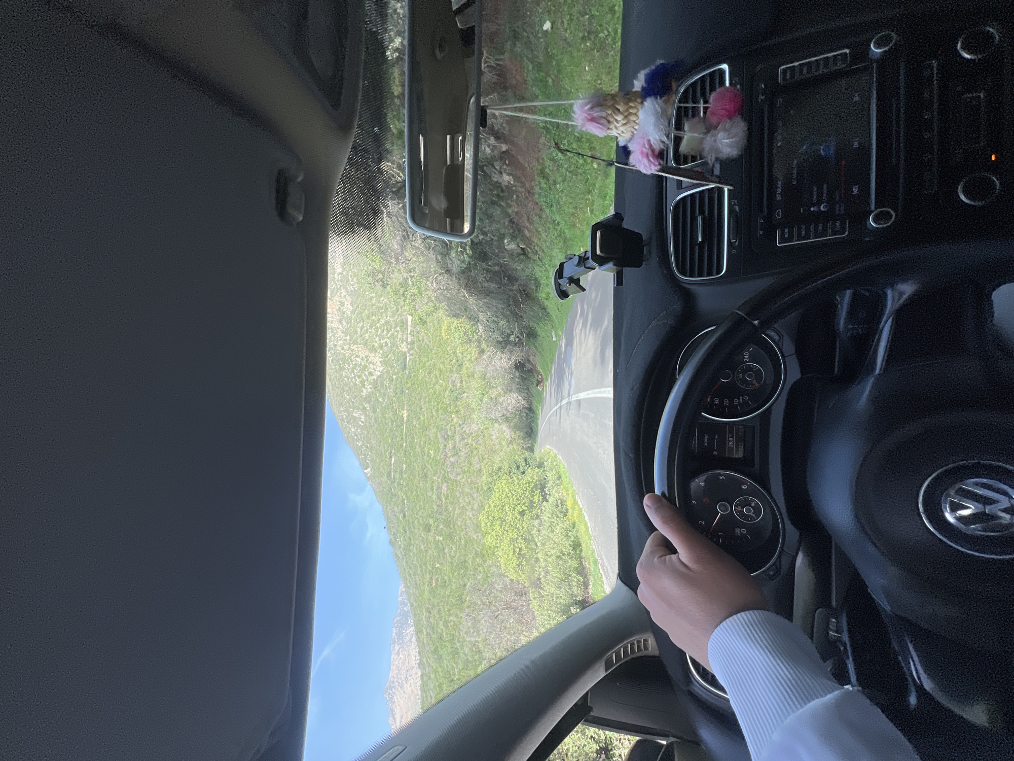 The view from inside a moving car shows a winding road leading through a mountainous, green landscape under a blue sky. The car's dashboard, steering wheel with a Volkswagen logo, and a hand holding the steering wheel are visible. A rearview mirror hangs from the roof, and a phone mount and decorative items adorn the dashboard. This is Foum Oudi, Morocco.