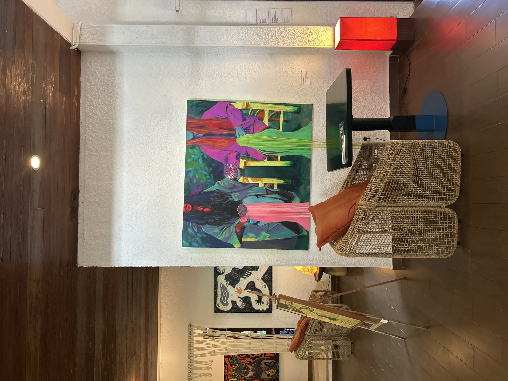 The image depicts an indoor art exhibition space, likely a gallery or cafe, in Mengwi, Indonesia. The room features a white, textured wall and a dark wood-paneled ceiling. On the wall are several paintings. A large, vibrant painting of two figures in colorful robes is prominently displayed, with a small label placed below it. To the left, another painting of a mythological creature is visible, and further left, a tapestry hangs. In the foreground, two wicker chairs with orange cushions are arranged. One chair is occupied by an artwork on an easel. A small, dark table with a blue base stands next to the other chair, and on it rests a book. A tall, red lamp illuminates the corner of the room, casting a warm glow. The floor is made of dark wood planks. The overall atmosphere suggests a contemporary and art-focused environment.