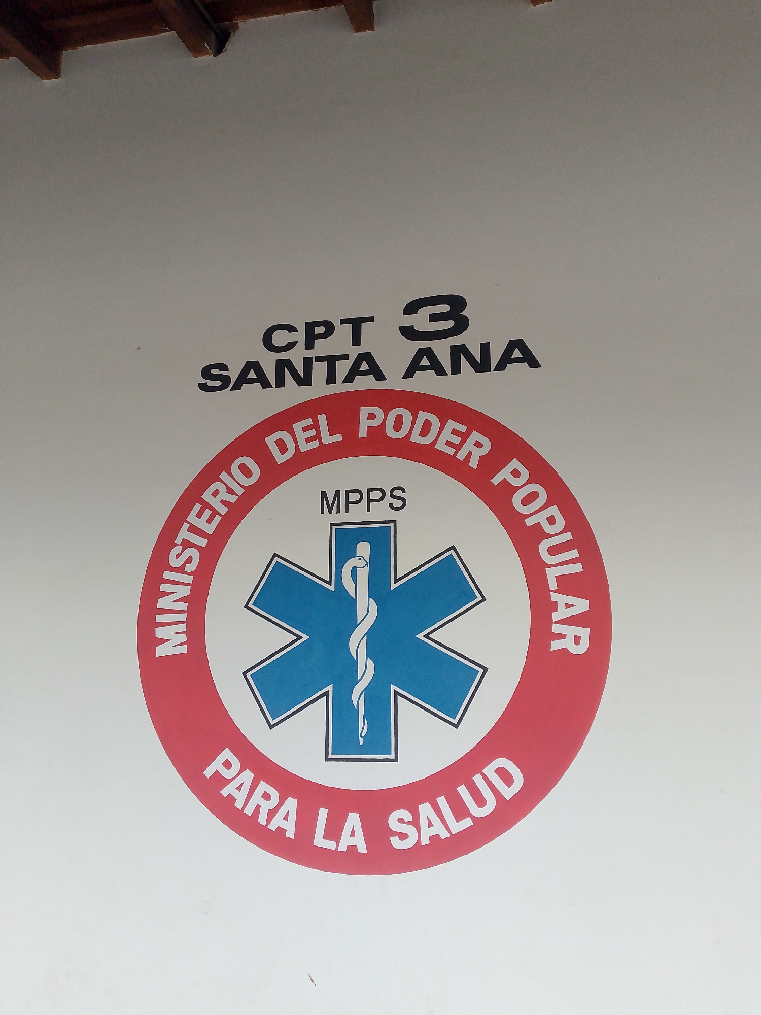 The image displays a prominent logo and text painted in black, red, and blue on a plain white wall, indicative of a governmental health facility in Santa Ana, Venezuela.
At the top, in bold black capital letters, are the designations "CPT 3" followed by "SANTA ANA". Below this, a large circular logo dominates the center. This logo features a wide red outer band, within which white capital letters read "MINISTERIO DEL PODER POPULAR" along the upper arc and "PARA LA SALUD" along the lower arc. The inner part of the circle contains a blue six-pointed Star of Life emblem, outlined in white. Inside the Star of Life, a white Rod of Asclepius is depicted. Directly above the Star of Life, within the logo, the acronym "MPPS" is written in smaller black capital letters.
The text "Ministerio del Poder Popular para la Salud" translates to "Ministry of Popular Power for Health," identifying the official health ministry of Venezuela. "MPPS" is the acronym for this ministry. The "CPT 3" likely refers to a specific primary care or triage center (Centro Primario de Triage) within the Santa Ana region. The Rod of Asclepius and the Star of Life are internationally recognized symbols for medicine and emergency medical services, respectively. The top edge of the image provides a glimpse of dark wooden architectural elements, possibly rafters or beams from an overhang or ceiling. No people are visible, and no active event is taking place; the image is a static shot of the wall signage.