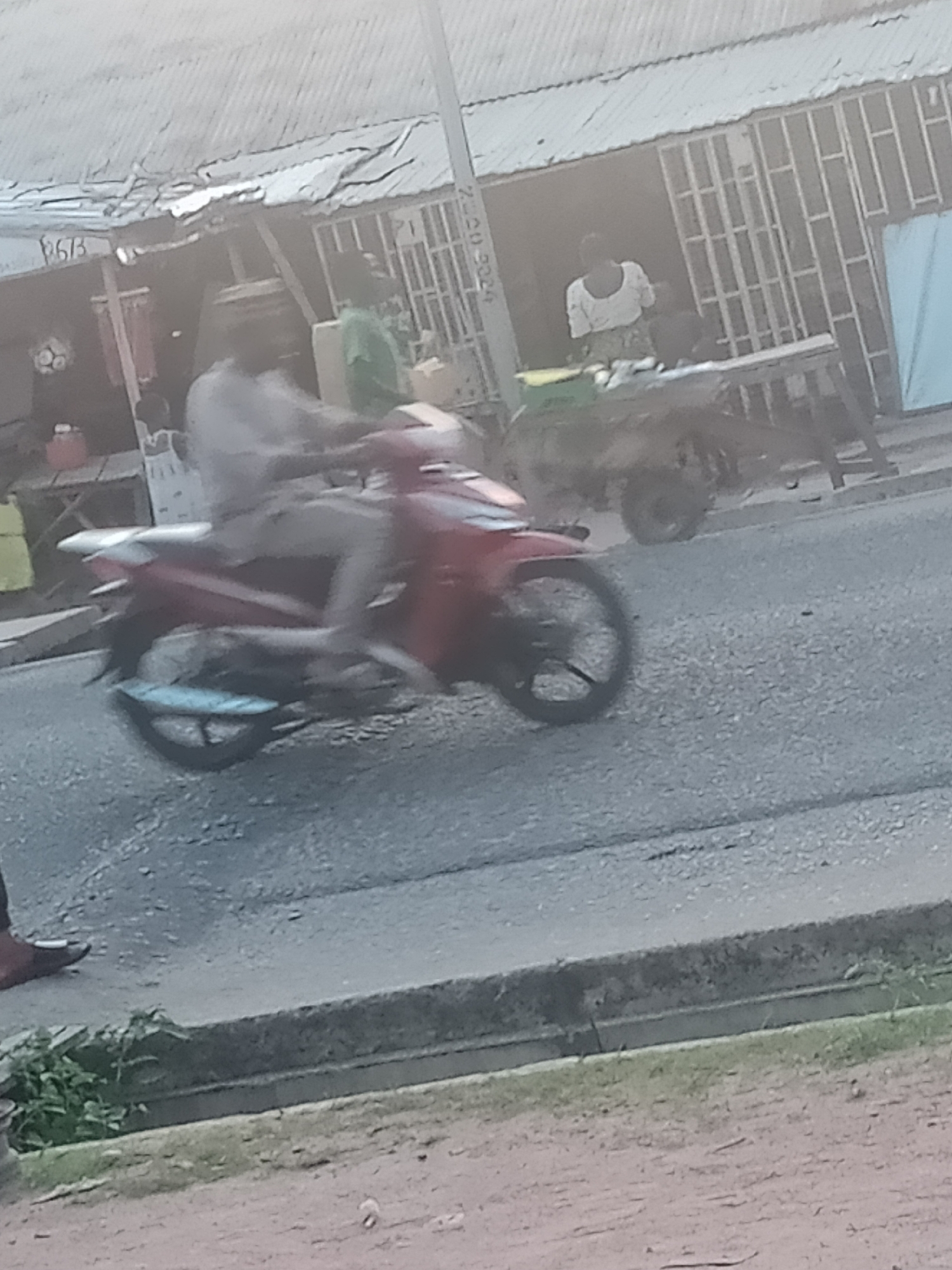 A motorcyclist rides a red and silver motorcycle in Jalingo, Nigeria. The motorcyclist is wearing a light-colored shirt and dark pants. The motorcycle is in motion, indicated by the blur. In the background, there is a building with a corrugated metal roof and barred windows, and a woman wearing a white shirt and patterned skirt stands near an open doorway. A cart with a wheel is visible to the right of the building. The road is paved with asphalt and there is a curb on the right side of the road with some greenery. The image is taken from a slightly low angle, capturing the lower half of the motorcycle and the road.