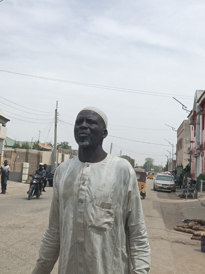 An outdoor scene in Hotoro South, Nigeria, depicts a dark-skinned adult male in the foreground, positioned slightly right of center, looking upwards and to his left. He wears a light-colored, vertically striped tunic with a chest pocket and four visible buttons, along with a light-colored skullcap. He has a short, patchy grey beard. The subject stands on an unpaved road.
In the midground, a two-person motorcycle travels away from the viewer on the left side of the road. Further left, a male in a white shirt walks. A building under construction with exposed concrete blocks and a green-roofed structure are visible beyond. Utility poles with overhead wires cross the upper portion of the frame.
The right side of the road extends into the distance, lined with buildings featuring red accents and external air conditioning units. Several individuals are visible sitting or standing along the street. A pile of large root vegetables is present on the ground near the right edge. Vehicles on the road include a yellow auto-rickshaw and a silver sedan. Streetlights are mounted on poles along the thoroughfare. The sky is overcast or hazy.