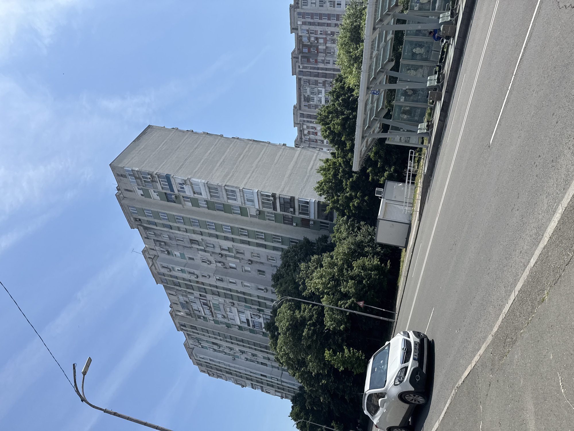 The image captures an urban street scene in Varna, Bulgaria, on a clear, sunny day. The dominant feature is a very tall, narrow, multi-story residential building (approximately 15-20 stories) constructed from grey and light green concrete panels, heavily adorned with numerous external air conditioning units and small balconies. In the background to the right, a shorter, multi-story residential building with grey and reddish-brown facades is visible.
In the foreground, a paved asphalt road extends diagonally from left to right, featuring double solid white lines in the center and a single dashed white line. A white first-generation Opel Mokka SUV, with Bulgarian license plate "A 8389 MC," is positioned in the left lane, appearing to move towards the viewer.
On the right side of the road, a modern bus stop with a translucent roof (likely glass or polycarbonate) and metal supports is present. Several metal benches are installed, and one person, a man wearing a blue top, is seated on a bench at the far end of the stop. The back panels of the bus stop display large posters. A blue circular sign featuring a wheelchair symbol is affixed to a support pole. A white utility box is situated behind the bus stop.
Lush, dense green trees line both sides of the road, partially obscuring the lower levels of the residential buildings. A tall street lamp with a single luminaire curves into the frame from the far left. A black utility wire stretches diagonally across the upper left portion of the bright blue sky, which contains sparse, wispy white clouds. A small, red-bordered triangular warning sign on a pole is discernible among the trees.