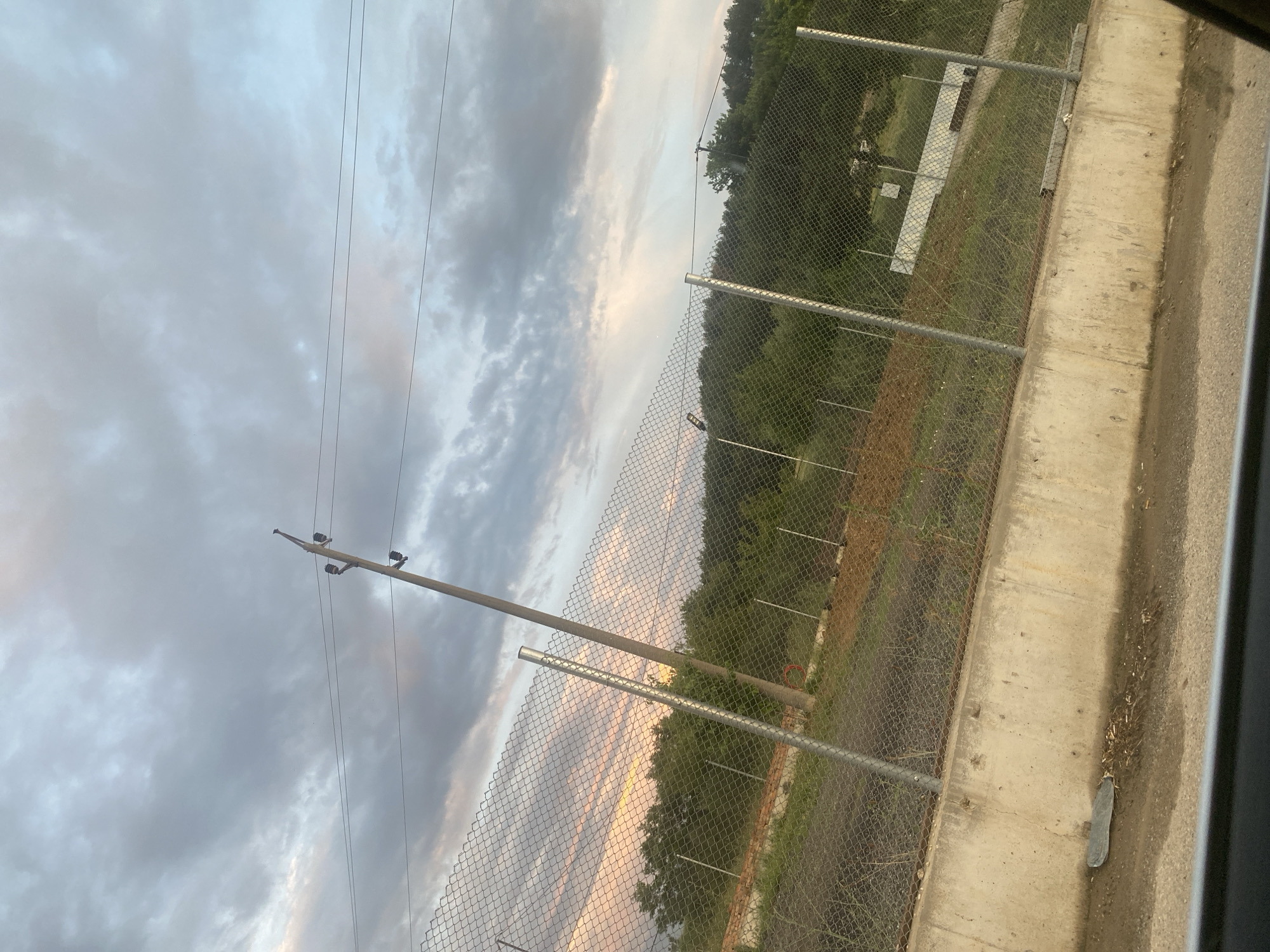 The image shows a cloudy sky with a sunset casting pink and yellow hues over the horizon. In the foreground, a chain-link fence runs horizontally across the frame, supported by metal posts. Behind the fence, there is a concrete retaining wall, and beyond that, a gently sloping hill covered in green trees. A wooden utility pole with insulators and wires stands diagonally against the sky. The scene appears to be on the outskirts of Ablanitsa, Bulgaria, possibly near a road given the view from a vehicle. There are no people or animals visible in the image. The overall impression is one of a rural or semi-rural landscape at dusk.