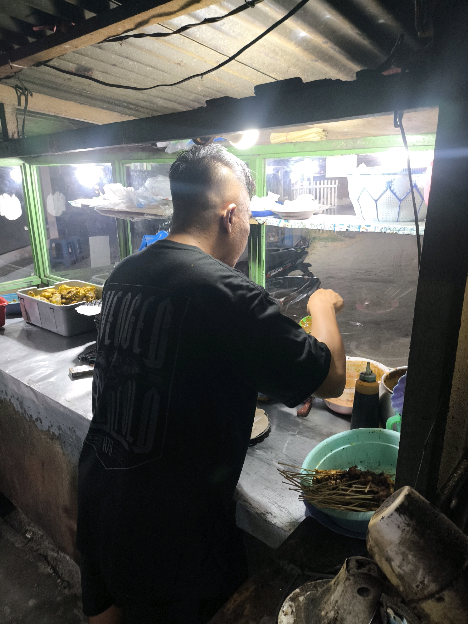 This image captures a street food stall at night in Luwoo, Indonesia. The stall is illuminated by overhead lights, casting a warm glow on the scene. A person, seen from the back, is standing behind a counter, preparing food. They are wearing a black t-shirt with a graphic design on the back. In front of them, on the counter, is a large container of cooked food, possibly satay, in a green bowl. Other items on the counter include bottles of sauce and serving utensils. The background shows the interior of the stall, with shelves holding various items, and through a window, a dimly lit outdoor scene with a motorbike visible. The overall environment suggests a bustling, yet humble, night-time food vendor.