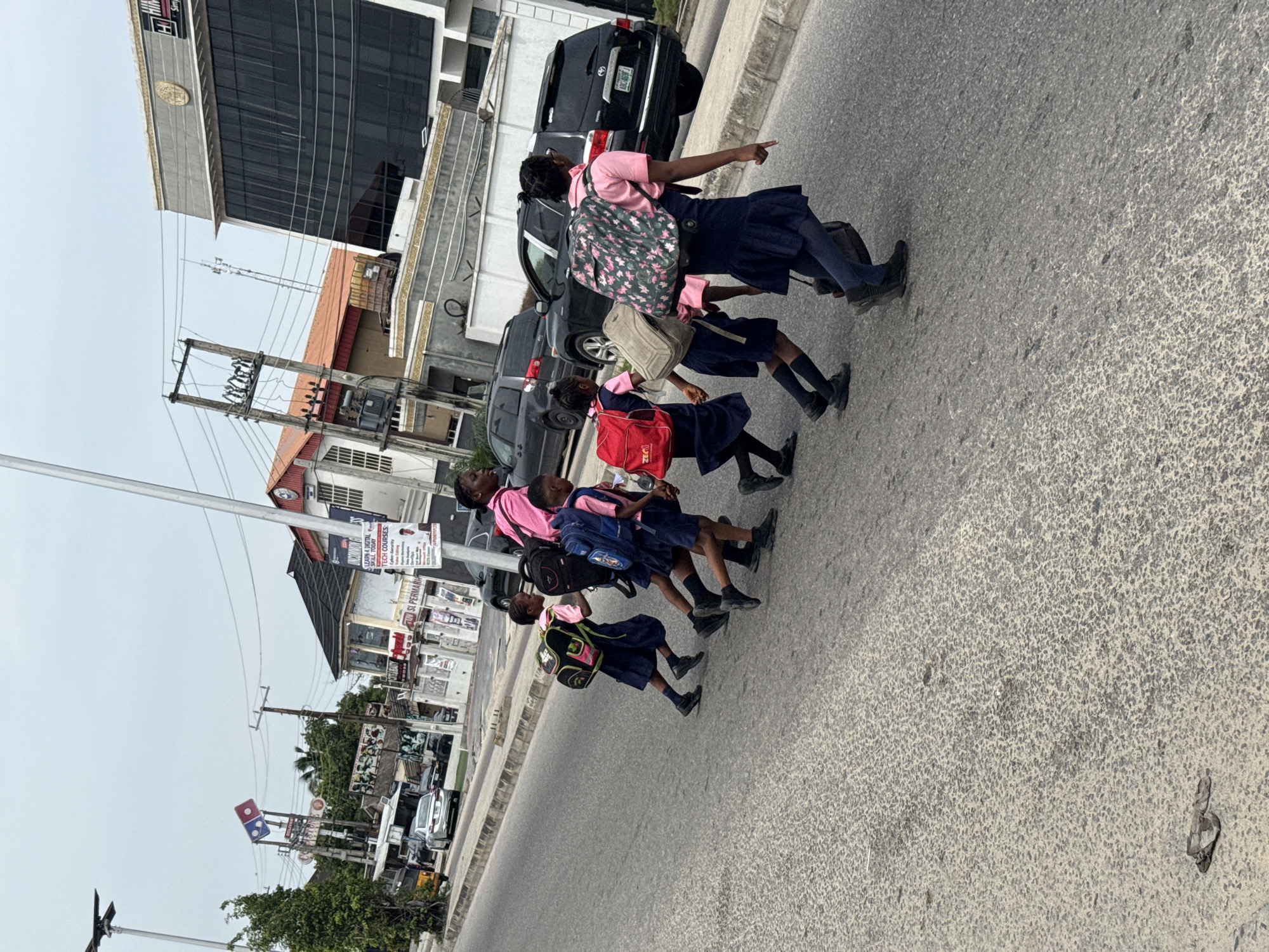 In Ado, Nigeria, a group of school children, likely uniformed in pink shirts and navy skirts, is crossing a paved road. They are carrying backpacks of various colors. An adult, also wearing a pink shirt, is leading them and pointing towards a building on the other side of the street. Several vehicles, including a black SUV, are parked or moving nearby. The background shows a mix of commercial buildings with signage and advertisements, power lines, and street lights.