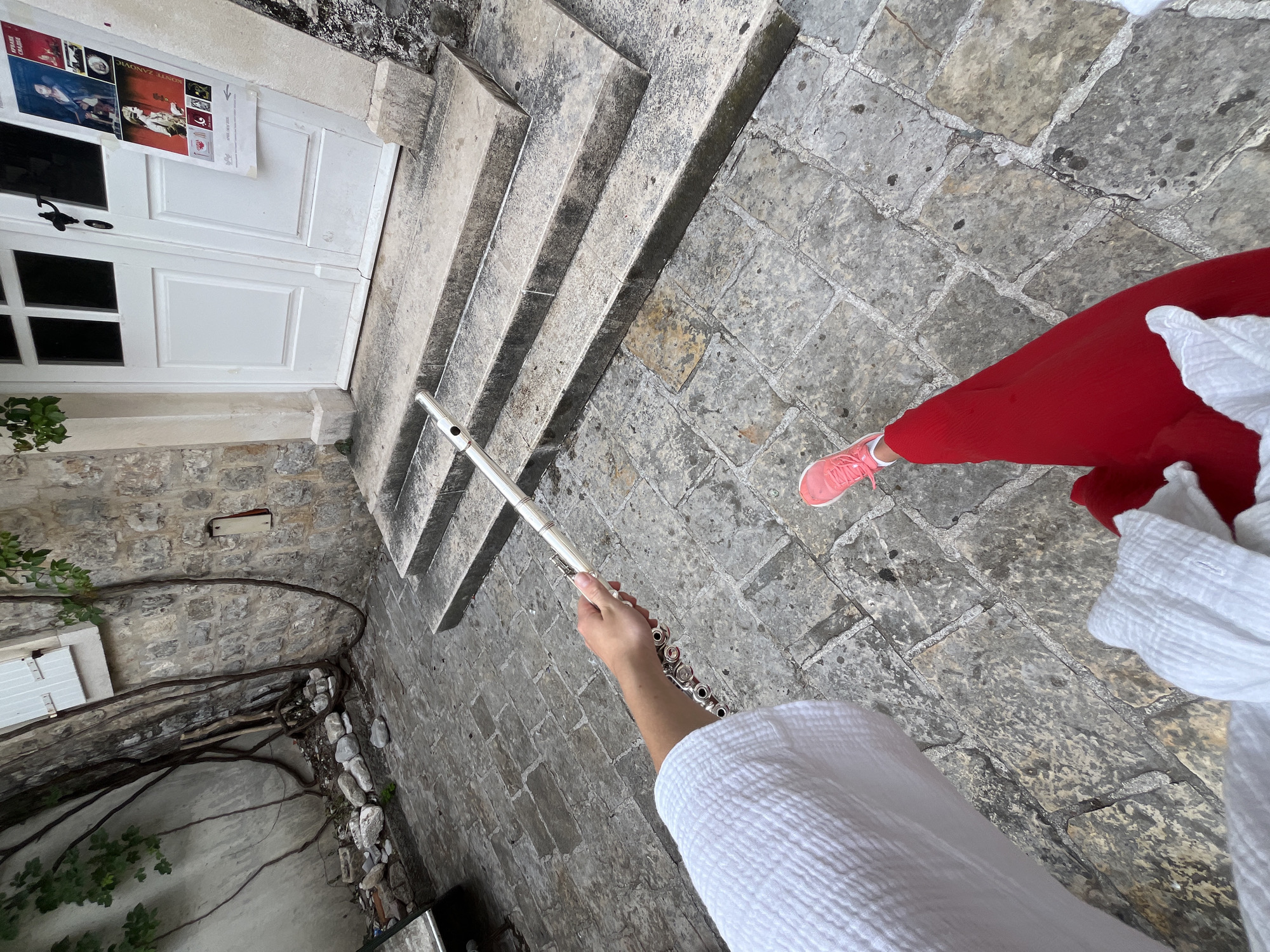 The image shows a person in a white robe and red pants holding a silver flute. The person is standing on a stone-paved surface near a set of stairs leading to a white door with posters on it. The location appears to be an old building, possibly in the city of Budva, Montenegro, given the architectural style and stone materials. There is also a wall covered with vines and a small window with white shutters on the left side of the frame. A pink shoe is visible in the top-right corner of the image. The composition suggests a first-person point of view, capturing the person's activity of potentially playing the flute in this location.