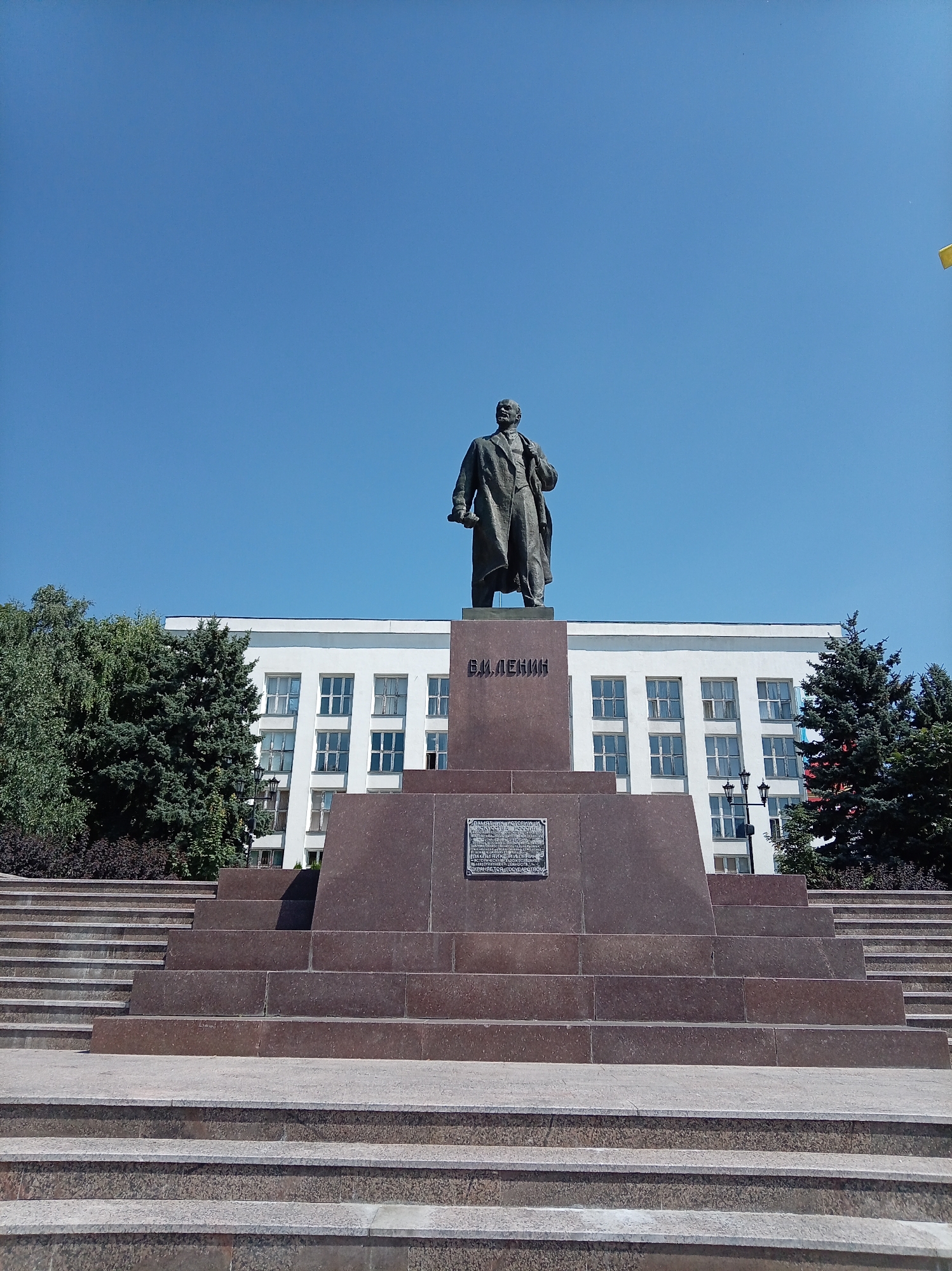 The image shows a statue of Vladimir Lenin in the city of Zheleznovodsk, Russia. The statue stands on a large granite pedestal in front of a white building with many windows. Trees surround the monument. The sky is clear and blue. The photo is taken from a low angle, looking up at the statue.