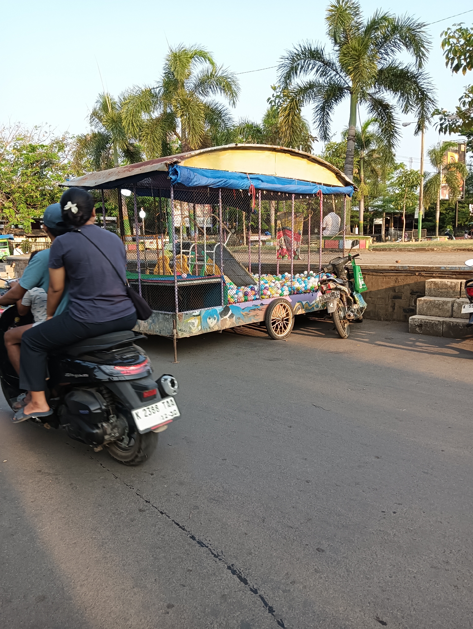 A black motorcycle is carrying two people and a trailer filled with colorful balls. The motorcycle is on a paved road in the city of Tayu, Indonesia. The trailer has a blue canopy and a fence made of wire mesh. Palm trees are visible in the background, along with buildings and a few other motorcycles. The sky is clear and blue.