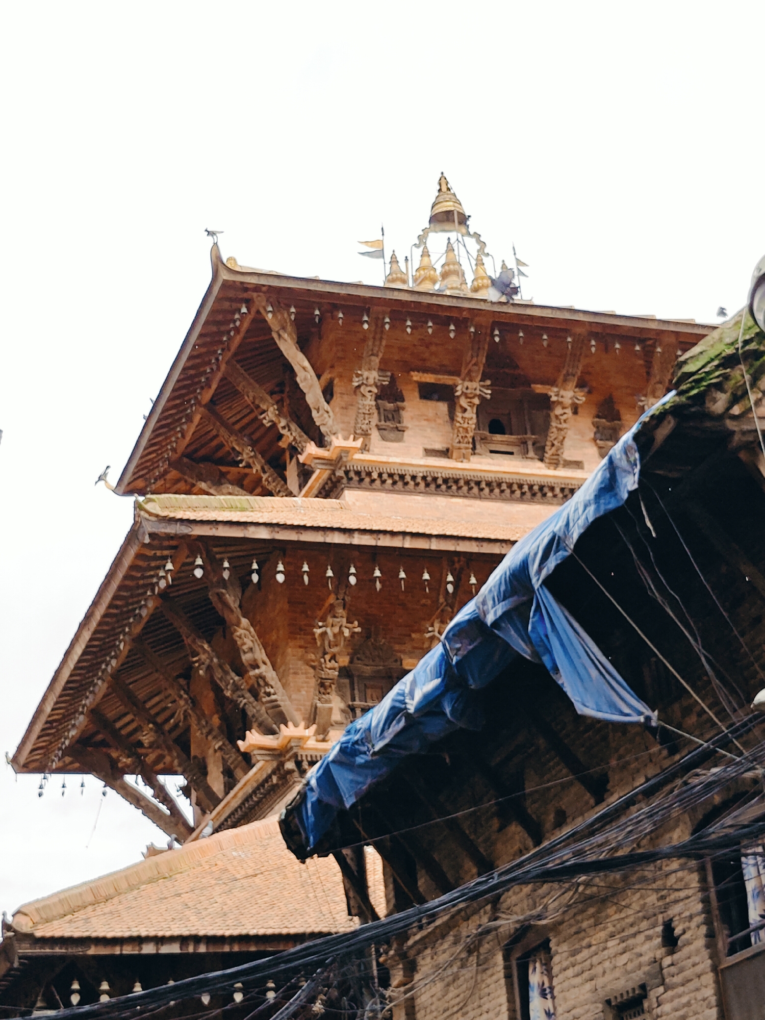 The image shows the upper portions of traditional wooden architecture, likely part of a temple complex in Lalitpur, Nepal. The structures feature multiple tiered roofs with intricate carvings and decorative elements, including small bells hanging from the eaves. A blue tarp is draped over a section of the foreground, obscuring part of the view and suggesting ongoing work or protection. Tangled electrical wires crisscross the lower portion of the frame, indicating the presence of modern infrastructure within the historic setting. The sky is overcast.