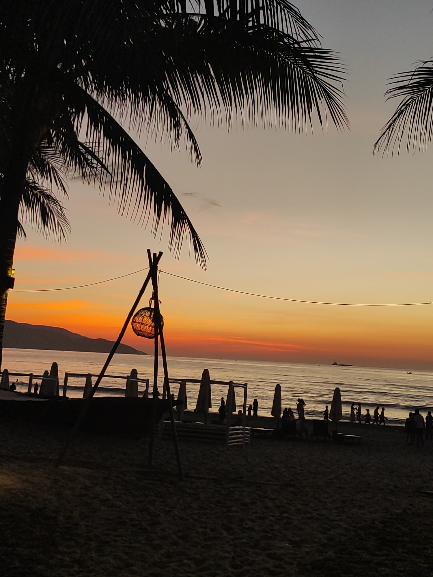 This is a photograph of a beach at sunset in Da Nang, Vietnam. The sky is a gradient of orange and pink, with a hint of purple at the top. The sun is setting behind a mountain range in the distance. The ocean is calm, with a few small boats visible on the horizon. In the foreground, there are palm trees silhouetted against the sky, and a beach umbrella is set up on the sand. There are also several people walking along the beach and sitting on lounge chairs. The overall mood of the photograph is peaceful and serene.