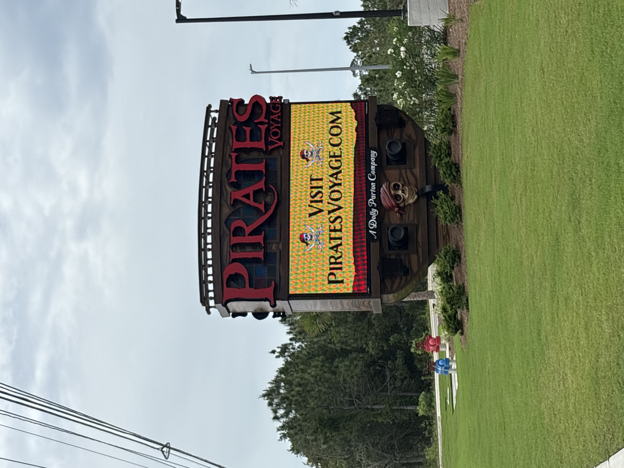 The image depicts a sign for "Pirates Voyage," located in Panama City Beach, Florida, USA. The sign is designed to resemble the stern of a pirate ship, featuring large red letters spelling "PIRATES" on the top, with a digital display screen to the right, showcasing "VISIT PIRATESVOYAGE.COM." A skull with a red bandana, and several cannons adorn the sign. The sign is situated beside a well-maintained green lawn, with trees and utility poles visible in the background.