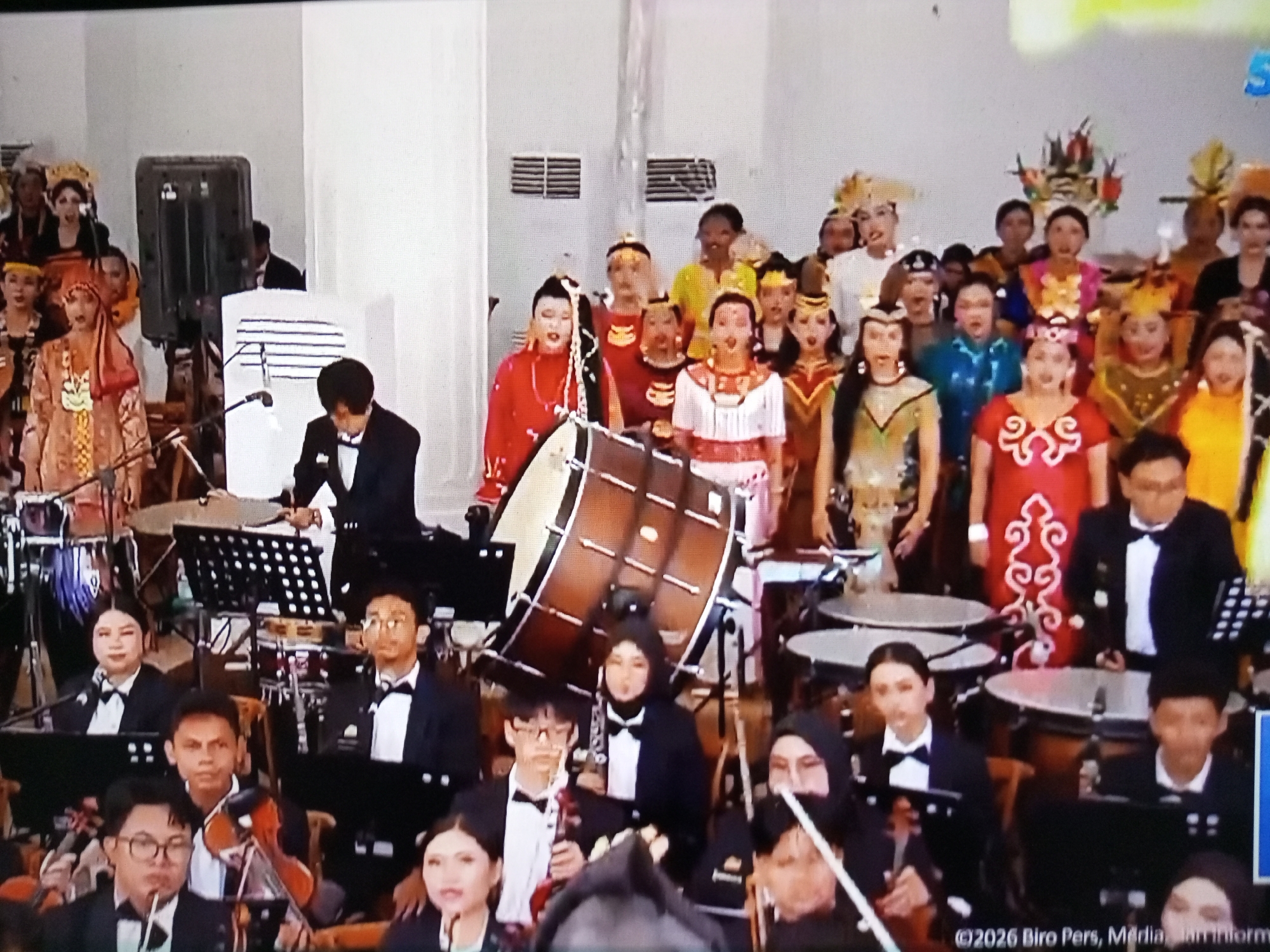 The image shows a musical performance, likely an orchestra, taking place indoors. A large drum is prominently featured in the center of the frame, with musicians seated behind it and to the sides. Several individuals in the foreground are playing instruments, including violins. Many of the performers are dressed in formal attire, with some also wearing traditional Indonesian costumes, suggesting a cultural event. A man in a suit is visible behind a music stand, possibly a conductor or presenter. The background features a white wall with some decorative elements and lighting. The overall atmosphere appears to be formal and celebratory. The location is identified as Banyudono, Indonesia, implying this event is taking place within that city.