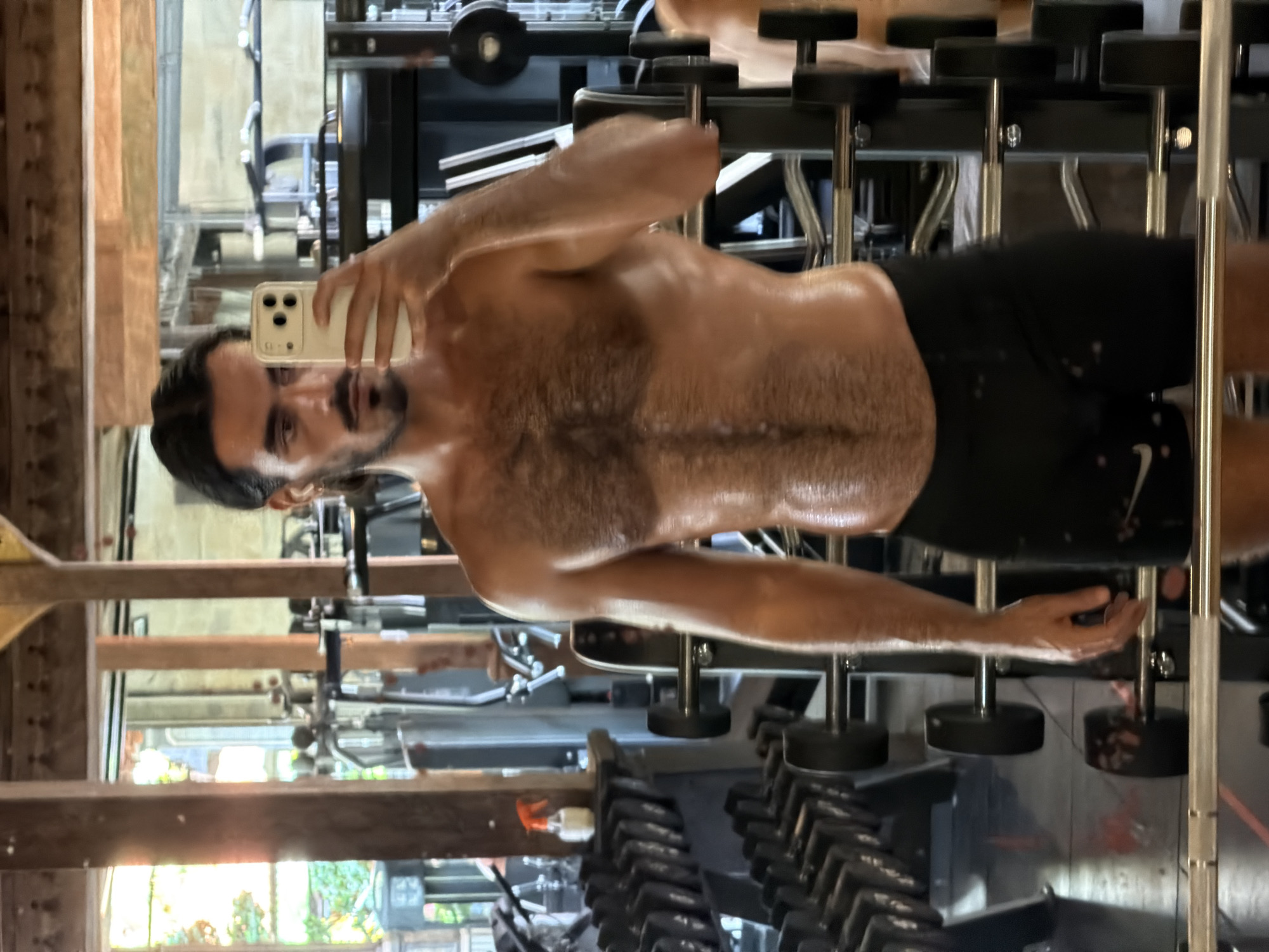 The image is an indoor shot of a man in a gym. The man is shirtless, wearing black shorts, and appears sweaty, possibly after a workout. He is holding a mobile phone, presumably taking a selfie in a mirror. Gym equipment, including dumbbells and a weight rack, are visible in the background. The location is possibly in North Kuta, Indonesia.