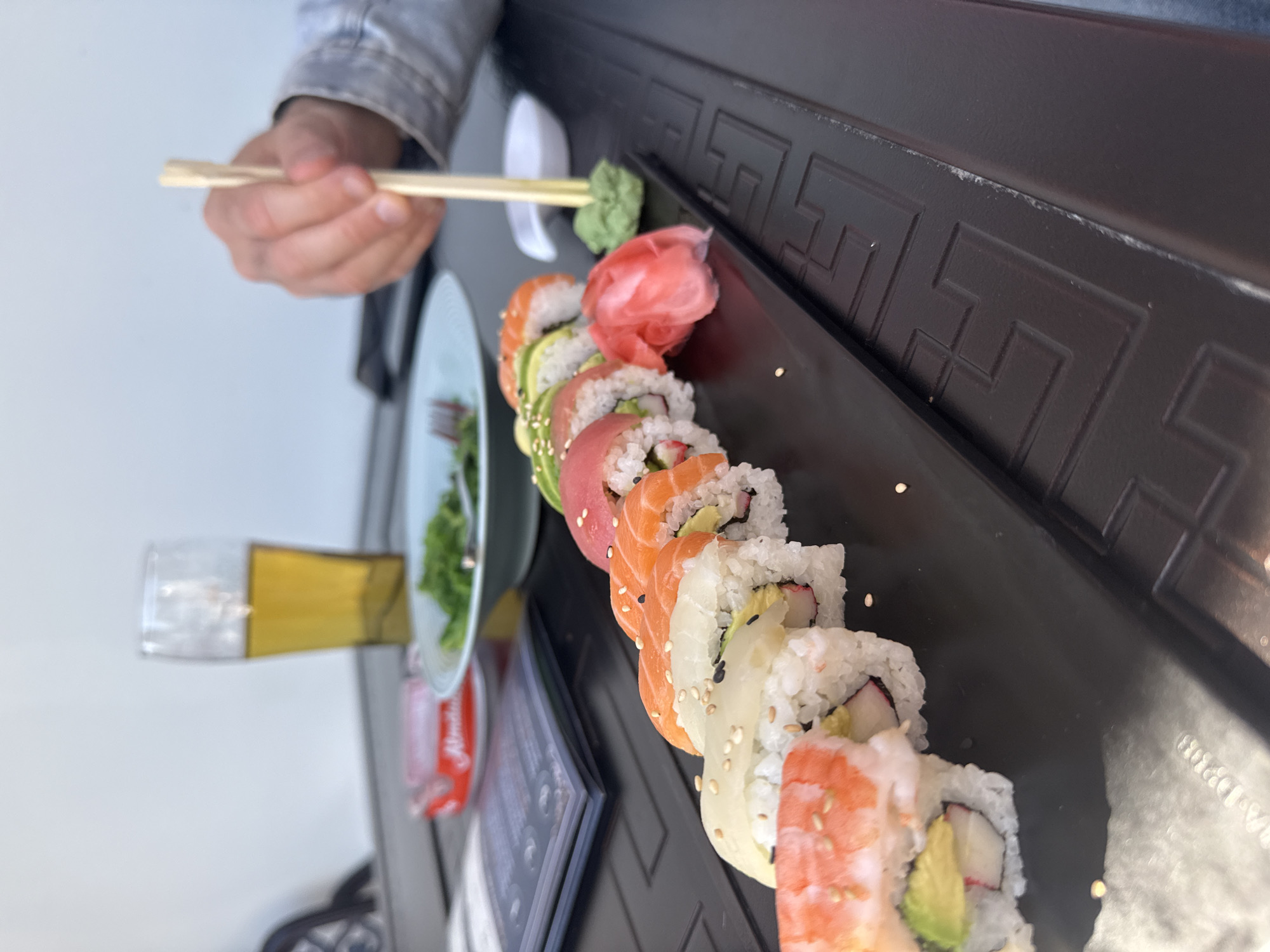 A close-up, eye-level shot captures a dining scene featuring a variety of sushi rolls on a dark, rectangular platter. The image is taken in what appears to be a restaurant or cafe in Hainburg an der Donau, Austria.
In the foreground, a person's hand, partially visible and wearing a light-colored denim jacket, holds a pair of wooden chopsticks. The chopsticks are poised over a small dollop of green wasabi, indicating the person is about to add it to their meal. Adjacent to the wasabi is a vibrant pile of pink pickled ginger (gari) and a white ceramic chopstick rest.
The sushi rolls are neatly arranged in a long line, showcasing different types, including maki rolls topped with fresh salmon, tuna, shrimp, and avocado, as well as rolls containing crab stick and sprinkled with sesame seeds. The dark platter has an embossed geometric pattern, possibly a Greek key design, running along its edge.
In the blurred background, a tall glass filled with a golden liquid, likely beer, is visible. To the left of the sushi plate, a light blue-grey plate holds a small portion of green salad and a fork. A red can with indistinct white text (possibly "Amstel" or a similar brand, though not clearly legible) and a dark menu or booklet are also present on the table. The overall impression is a casual yet appealing meal.