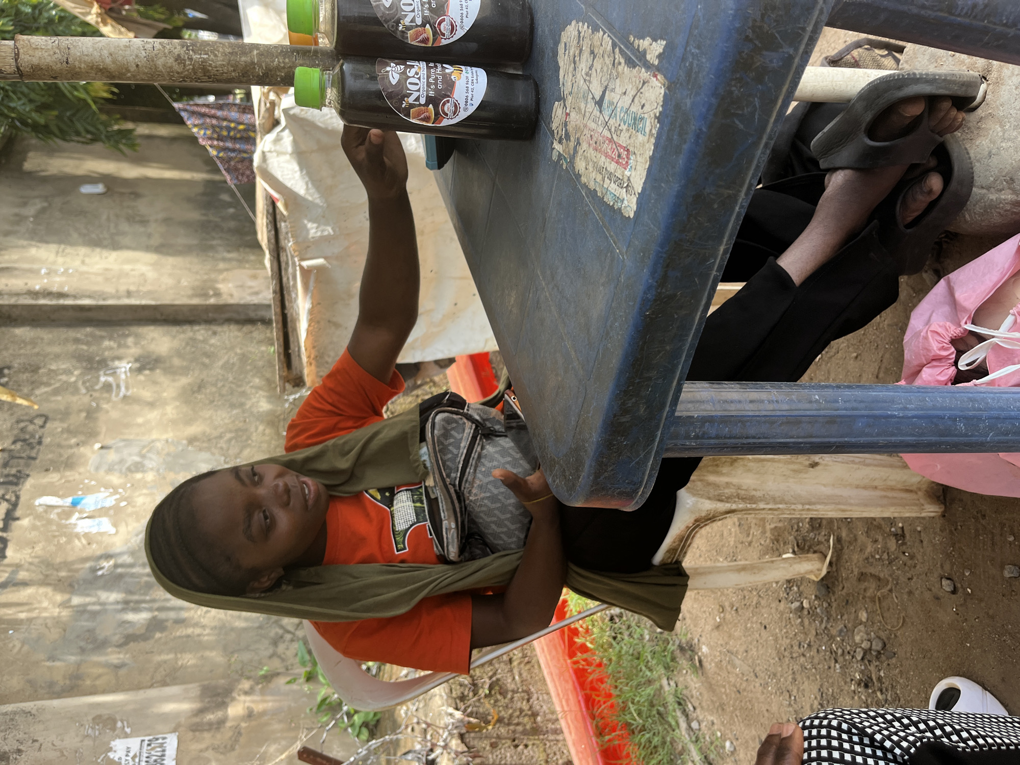 In an outdoor setting in Abuja, Nigeria, a young person with dark skin and braids sits at a blue plastic table. They wear an orange t-shirt, a green scarf draped around their head and shoulders, and black pants. A small, gray and black patterned bag is slung across their chest. Their feet, clad in black flip-flops, rest on the table. The person is looking towards the camera with a slight smile.
On the table, two glass bottles of what appears to be honey are visible, with labels that read "NOSH Organic and Natural" and "It's Pure, Rich and Healthy." The table has stickers with text, including "SY PAY" and a phone number. Behind the table, there's a concrete wall and some greenery. The lighting suggests it is daytime. The overall scene appears to be a casual outdoor market or roadside stall.
