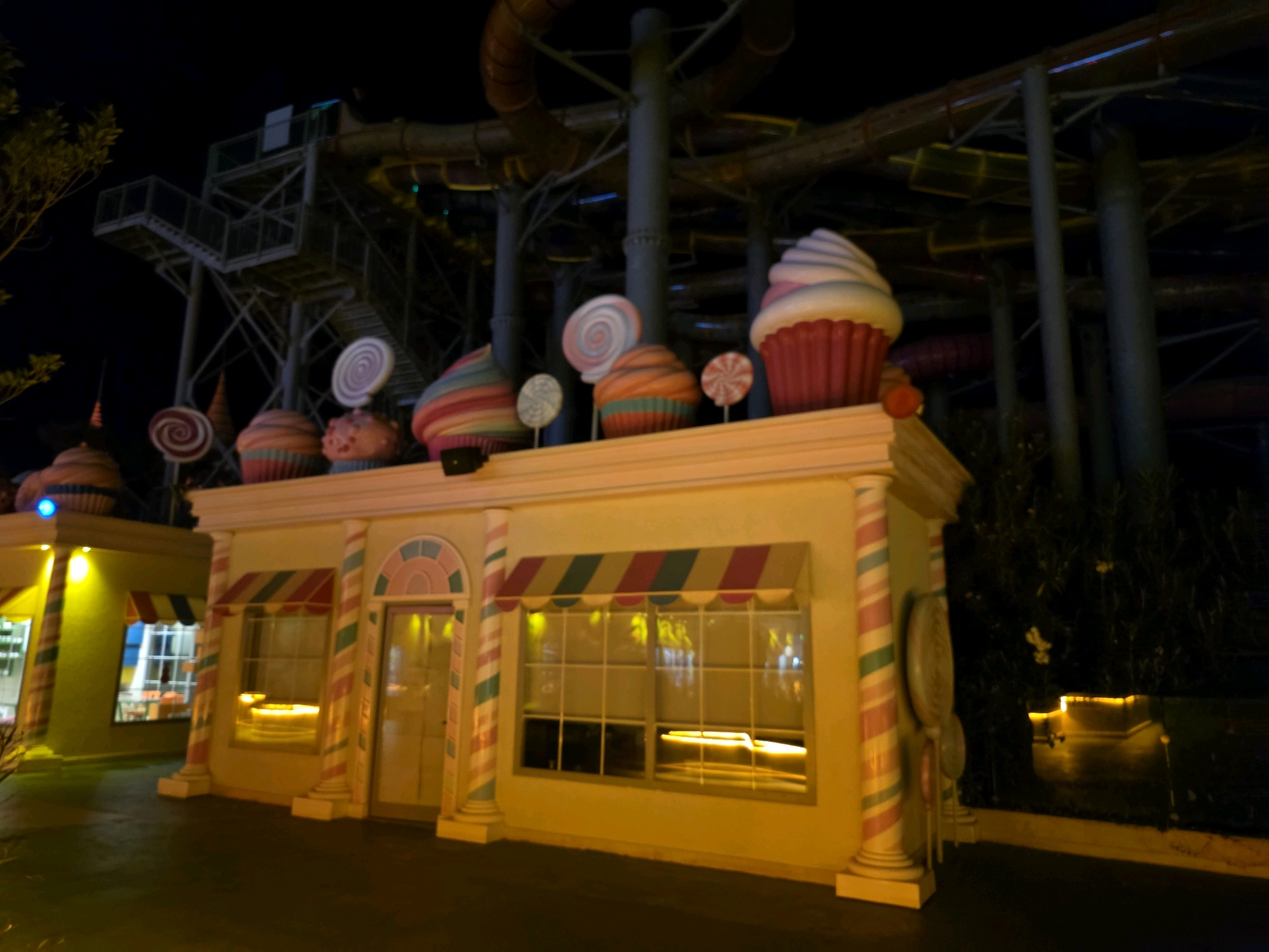 This is a nighttime exterior shot of a candy-themed attraction at the Dedeman Aquapark in Bodrum, Turkey. The foreground features a whimsical building with striped columns, awnings, and large decorative cupcakes and lollipops. The building appears to be a shop or entrance area, with lit windows and a glass door. In the background, a section of a large water slide structure is visible, illuminated by artificial lights. The overall scene evokes a playful and fantastical atmosphere, typical of a theme park or water park at night. There are no people visible in the image.