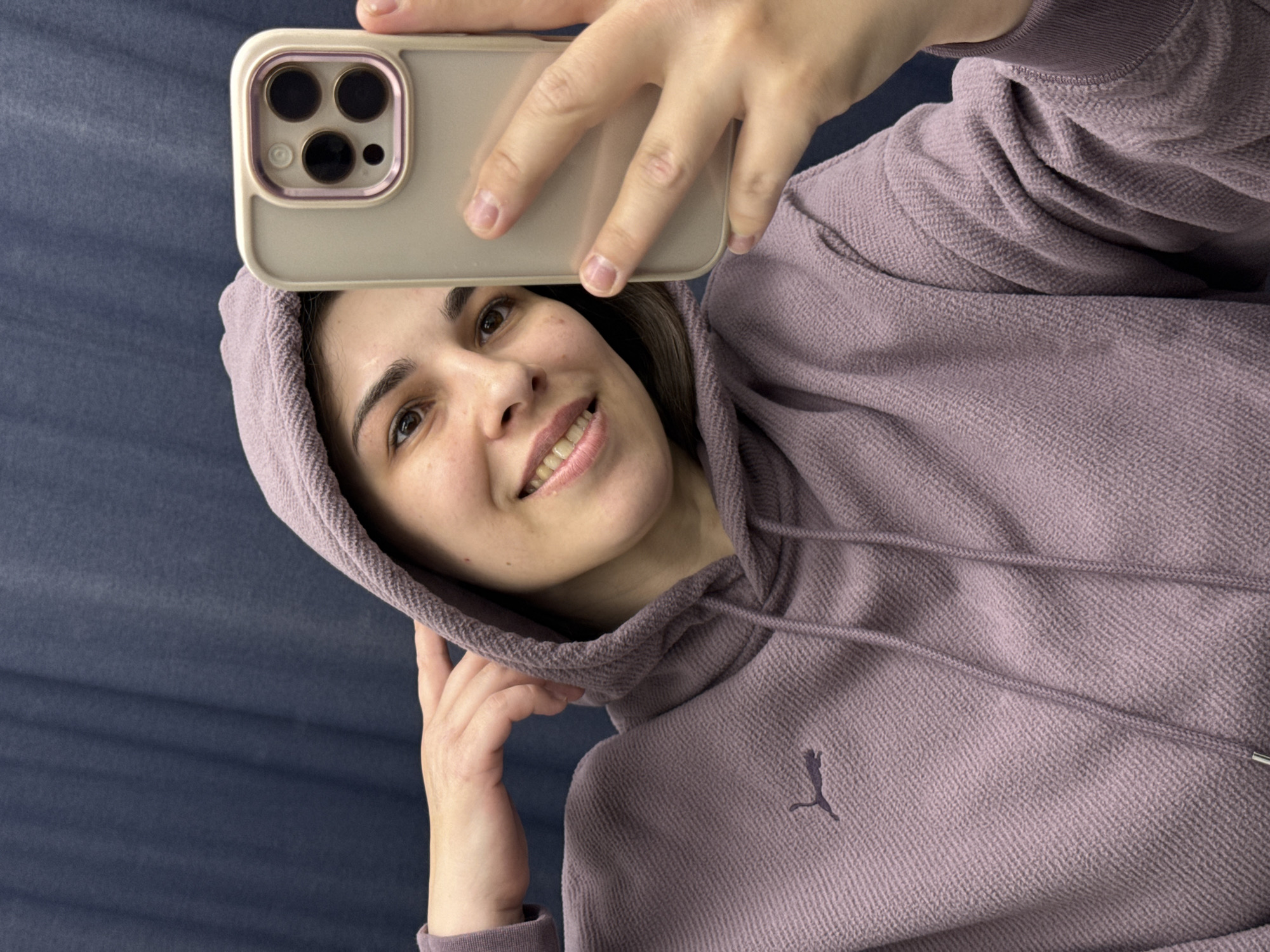 A woman in a purple Puma hoodie takes a selfie with her smartphone. The background is a plain blue. The location is not discernible from the given information.