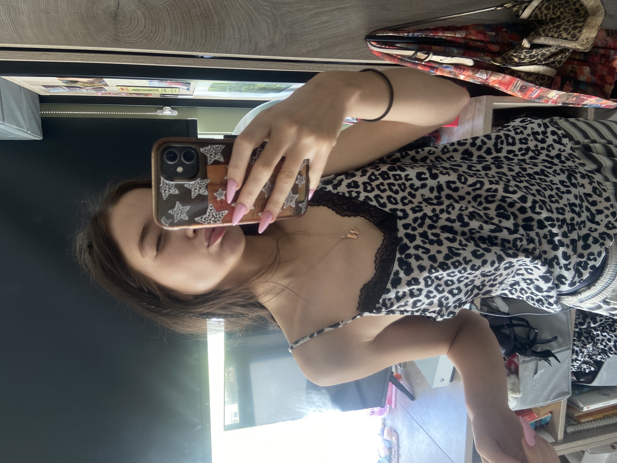 A woman is taking a selfie in what appears to be a bedroom. She is wearing a leopard print top with black lace detailing. She is holding a smartphone with a decorative case. The background includes a dresser, a television, and various items on shelves. The location is not possible to determine with the given information, so no location in London, United Kingdom, can be specified.