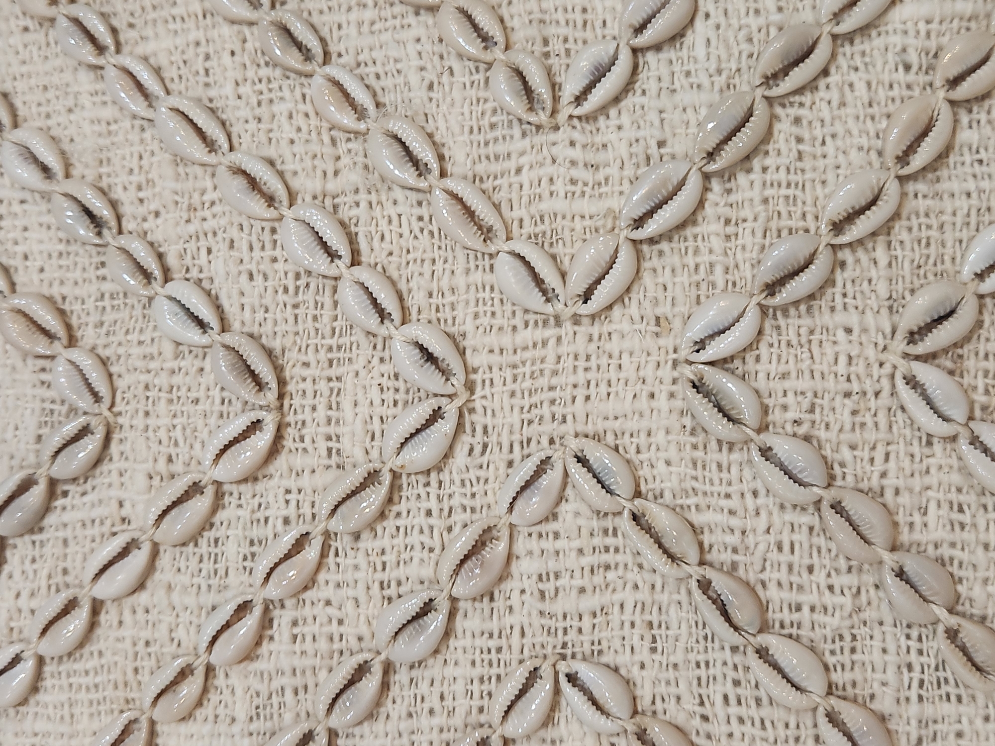 A close-up, overhead view of a natural fiber textile with a decorative pattern of small, white cowrie shells strung together on thin white thread. The shells are arranged in diagonal lines forming a diamond-like pattern across the fabric. The textile appears to be a loosely woven, textured material, possibly linen or a similar natural fiber, in a light beige or off-white color. The shells themselves are smooth and glossy, with a characteristic dark opening. There are no people or other objects visible in the image. The lighting is even, casting minimal shadows, highlighting the texture of both the shells and the fabric. The image is sharp and in focus, allowing for clear detail of the individual shells and their arrangement. The location in Maitland, United States of America, is not evident from the visual information present in the image.