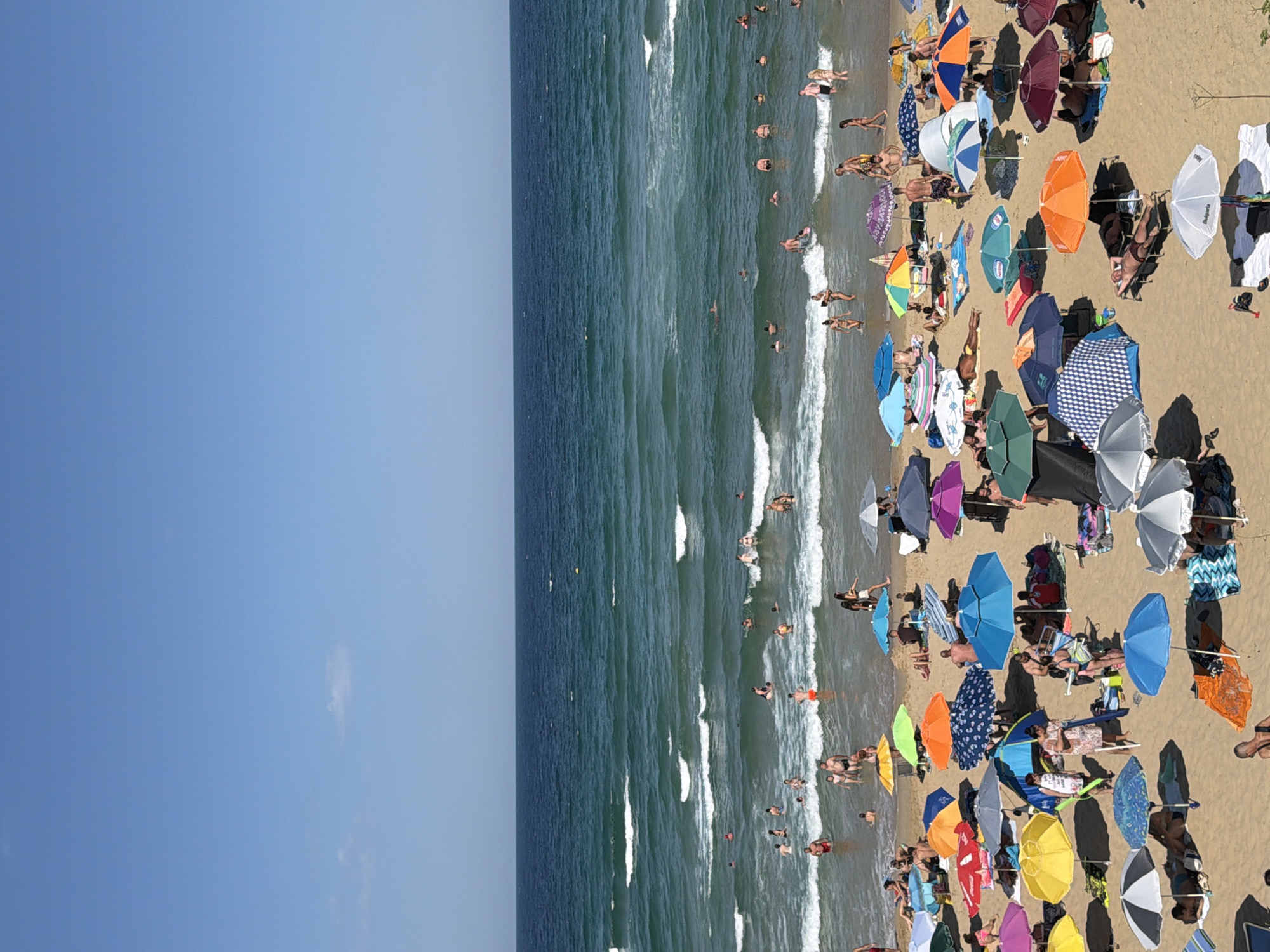The image depicts a crowded beach in Primorsko, Bulgaria, on a sunny day with a clear blue sky. The foreground is dominated by a sandy beach dotted with numerous colorful beach umbrellas and a few beach tents. Many people are seen relaxing under the umbrellas, sunbathing on towels, or sitting on beach chairs. Some individuals are walking on the sand, while others are in the process of setting up or packing their belongings.
The middle ground features the shallow waters of the sea, where a large number of people are swimming, wading, and playing in the gentle waves. Children are visible among the swimmers, indicating a family-friendly atmosphere. The sea itself is a vibrant turquoise color, with white foam from the waves breaking near the shore.
The background shows the deeper blue ocean stretching towards the horizon, meeting the clear blue sky. There are no boats or other vessels visible in the water. The overall scene conveys a sense of leisure and recreation, typical of a popular seaside resort during peak season.