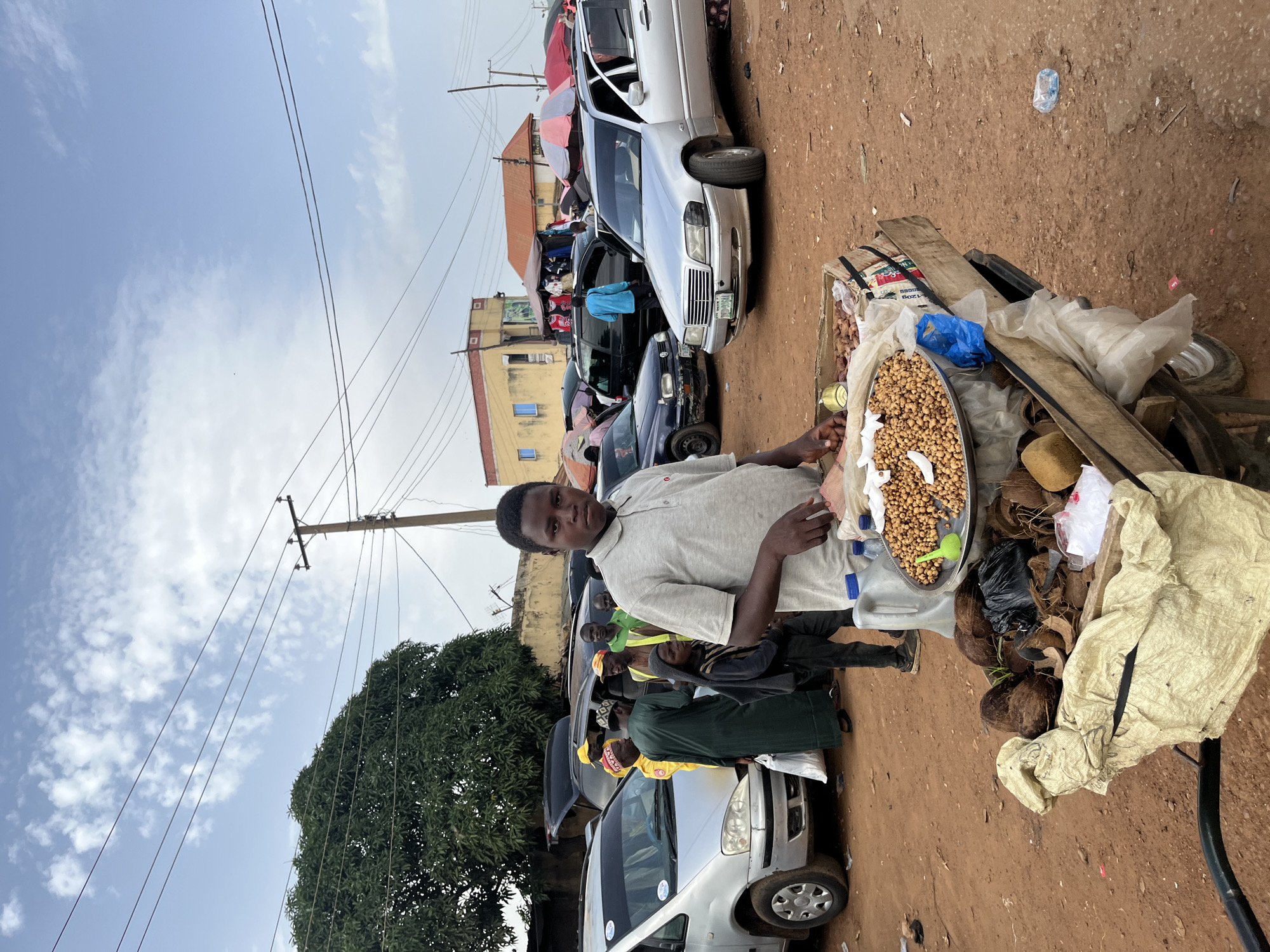 The image depicts a vibrant street scene in Rafin Par, Nigeria. In the foreground, a young man, likely a vendor, is standing next to a cart laden with goods. He is wearing a light gray polo shirt and appears to be looking directly at the camera. The cart displays a large metal tray filled with what appear to be nuts or seeds, accompanied by various other items like coconuts and bags of produce. The setting is a dusty, unpaved street, characteristic of many marketplaces in Nigeria.
Behind the vendor, several cars are parked along the roadside, suggesting a busy commercial area. Some of these vehicles are partially obscured by umbrellas, possibly indicating market stalls or shade for shoppers. In the background, buildings with yellow and red facades are visible, along with overhead power lines. The sky is partly cloudy, suggesting it could be mid-morning or afternoon. The overall atmosphere appears lively and bustling, with people engaged in daily commerce. The image captures a slice of everyday life in a Nigerian marketplace.