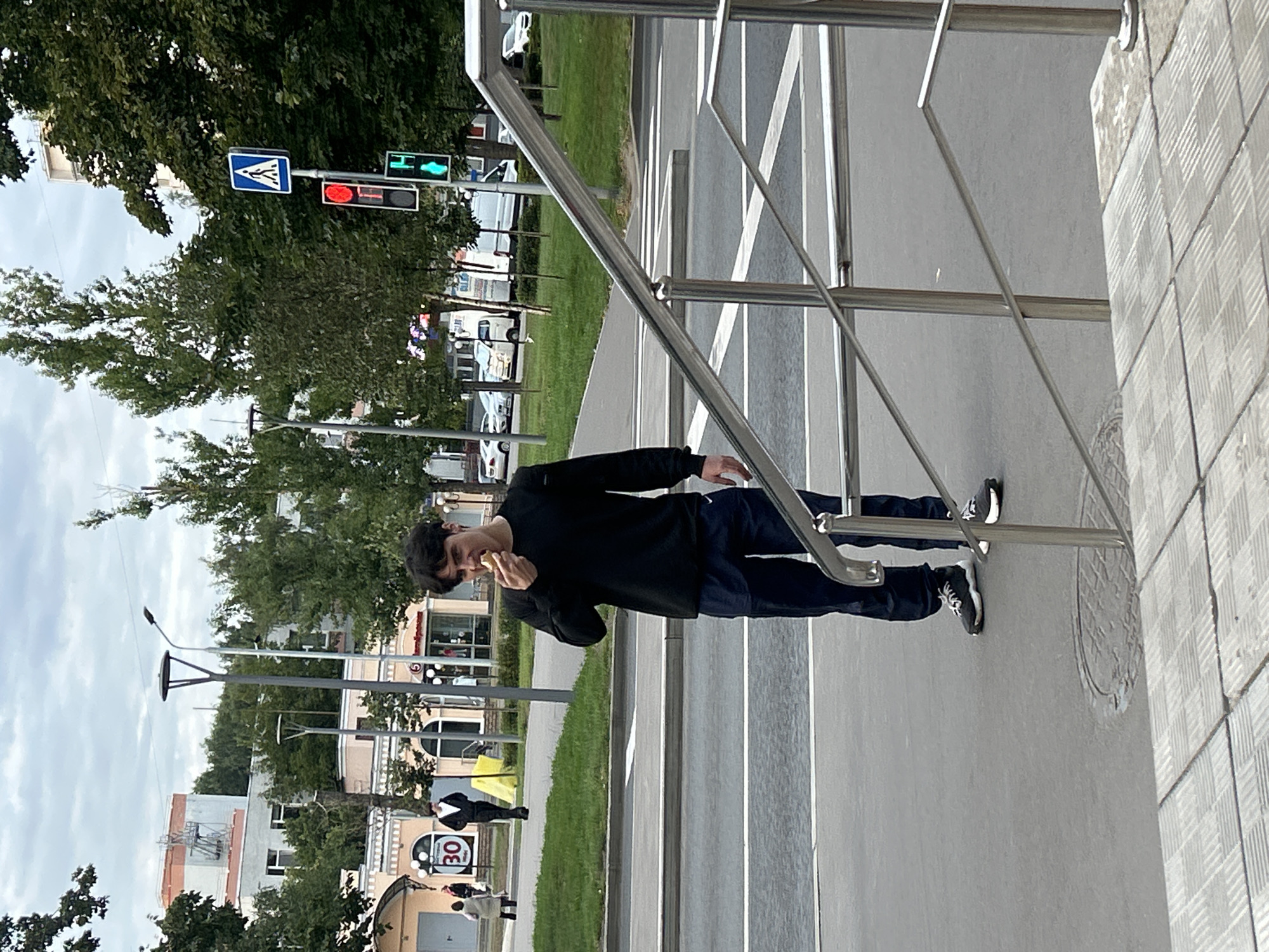 A young man with dark hair is standing on a sidewalk in Moscow, Russia, eating a piece of food. He is wearing a black long-sleeved shirt and dark blue pants. The man is facing the viewer and is in the process of taking a bite of food. In the background, there are trees, buildings, and traffic lights. A pedestrian crossing is visible on the street in front of the man. The man appears to be alone and is not interacting with anyone. The overall scene suggests a casual moment in an urban environment.