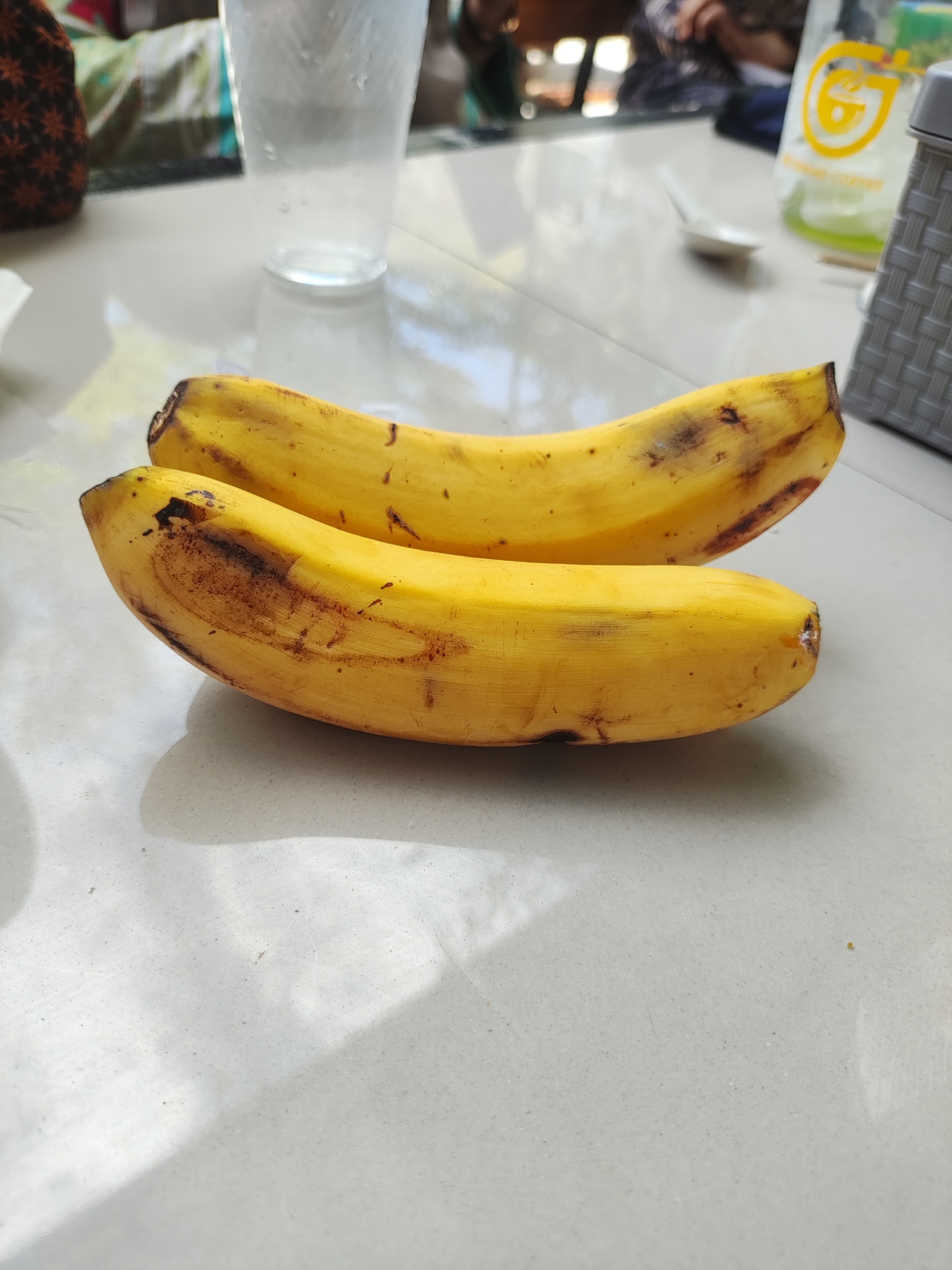 On a tiled surface, likely a table or countertop, are two ripe bananas with brown spots, indicative of ripening. A clear glass of water sits in the background. Other indistinct objects are visible, including a plastic container with a yellow logo and a gray woven container. There is no visible indication of the location being Lhokseumawe, Indonesia. No people are directly interacting with the bananas.