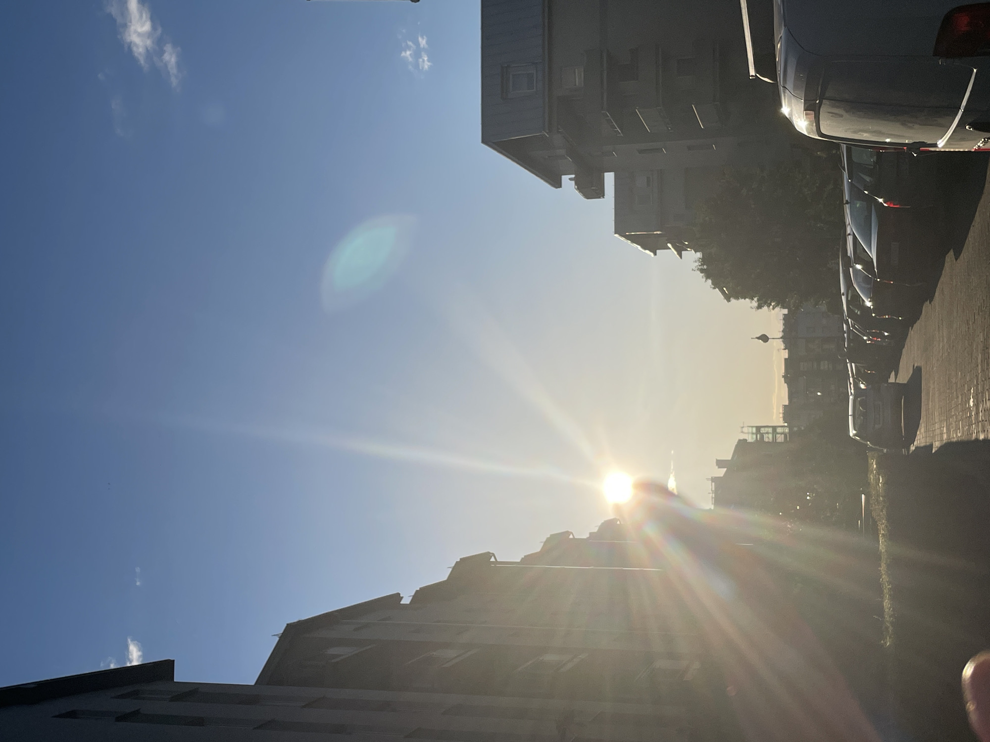 The image depicts an urban street scene at sunrise or sunset in the city of Unknown, Unknown. The sun, positioned low in the sky towards the left-center, casts intense light with pronounced lens flare, characterized by a white starburst effect and rainbow-colored rays emanating from its core. A diffuse, greenish-blue oval lens flare is also visible in the upper right quadrant of the otherwise clear, light blue sky, which contains a few sparse white clouds.
Multi-story residential buildings line both sides of the street, appearing largely in silhouette due to the backlighting. The buildings on the left are dark and show multiple windows and balconies. The buildings on the right feature distinct sloped roofs with dormer windows, also silhouetted. A row of dark-colored cars, including sedans and a light-colored van in the immediate foreground on the right, are parked facing away from the viewer along the street. The ground in the foreground on the left appears to be paved with pavers or cobblestones, bordering a low green hedge. Beyond the parked vehicles, trees with dark foliage are visible, and a distant lamppost or utility pole stands upright. No people are visible in the scene. A small, out-of-focus, reddish object (possibly a fingertip) is present in the bottom-left corner of the frame.