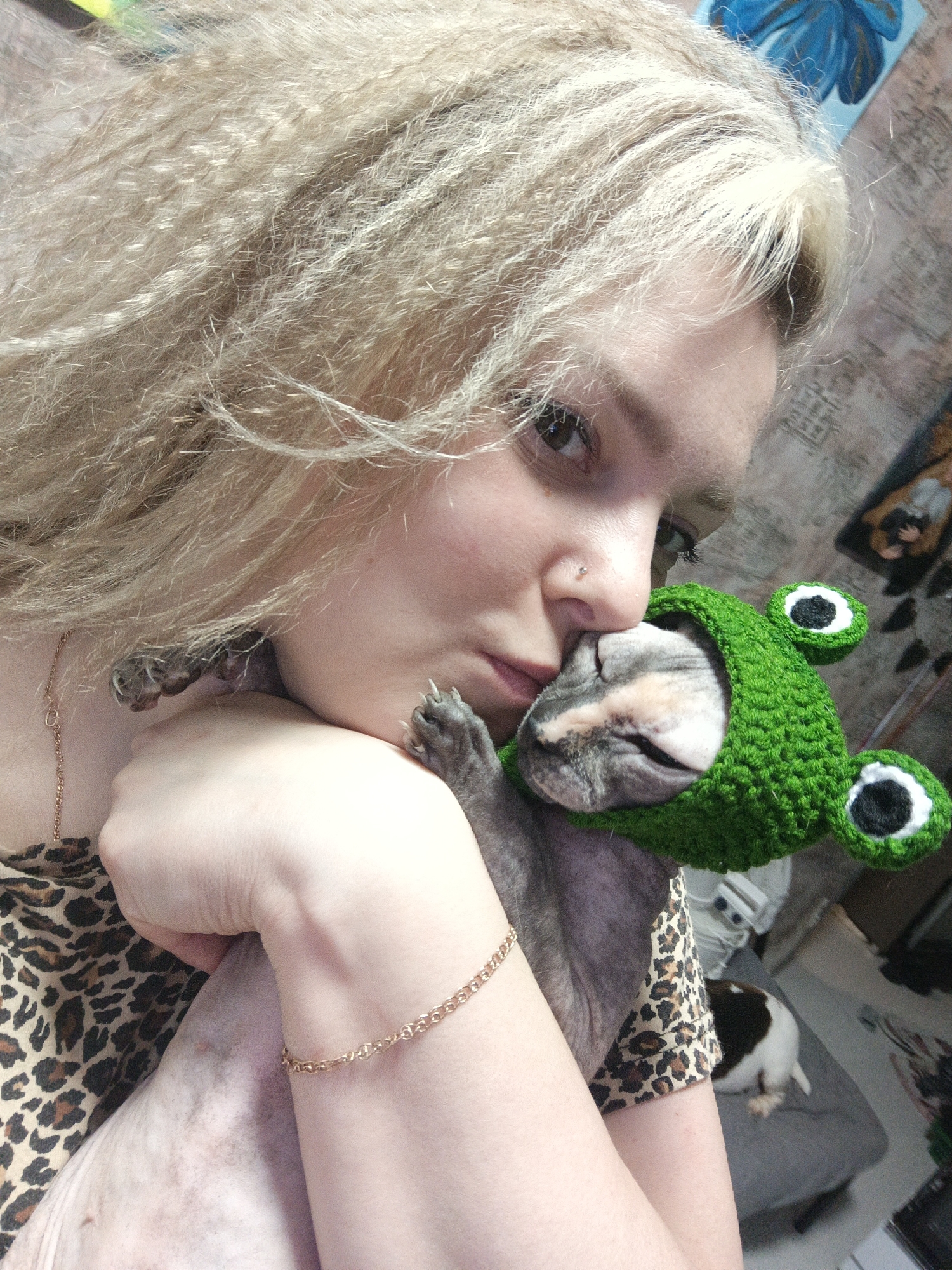 A close-up selfie captures a young woman tenderly interacting with her pet hairless cat, likely a Sphynx, indoors in Markovsky, Russia. The woman has light skin, blonde crimped hair, and light-colored eyes, with a small nose piercing. She is wearing a leopard print top, a delicate gold chain necklace, and a matching gold link bracelet on her left wrist.
She holds the cat close to her face, gently nuzzling its head with her nose and mouth, conveying a strong sense of affection. The cat, appearing calm and possibly sleepy, is distinctive in a vibrant green crocheted hat designed to look like a frog, complete with large black and white eyes. The cat's greyish, mottled skin is visible on its body and paws. Its tiny paws are seen resting against the woman's arm and hair.
The background is softly blurred but reveals elements of a casual indoor setting. A textured wall is visible behind the woman's head, and a small, abstract piece of artwork or decoration can be seen in the upper right corner. In the lower right background, another pet, a black and white cat or small dog, is partially visible resting on grey furniture. The lighting suggests daytime. The overall mood of the image is warm and intimate, highlighting the bond between the woman and her uniquely dressed pet.