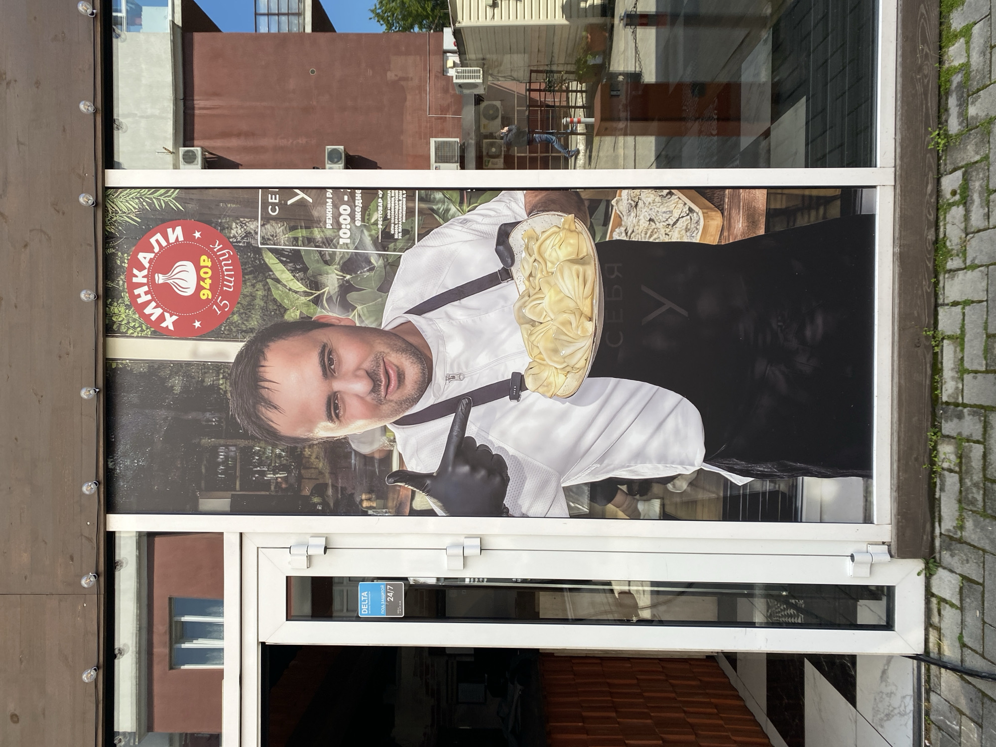 The image shows the entrance to a restaurant in Sochi, Russia. A large poster on the glass door features a man in a chef's uniform holding a plate of khinkali (Georgian dumplings). A red circle on the poster advertises "15 khinkali" for "940P" (presumably rubles). The restaurant's name includes "Khinkali." The poster also includes "REZHIM RA," indicating operating hours are "10:00-ezhedne," (10:00-daily). The doorframe is white, with a small blue and white sticker on the lower pane which reads "DELTA POD ZASHCHITOI 24/7" (DELTA Under Protection 24/7). The building façade is a combination of brown wooden panels and plaster. The surrounding environment appears to be a street scene with other buildings visible in the reflection on the glass door.