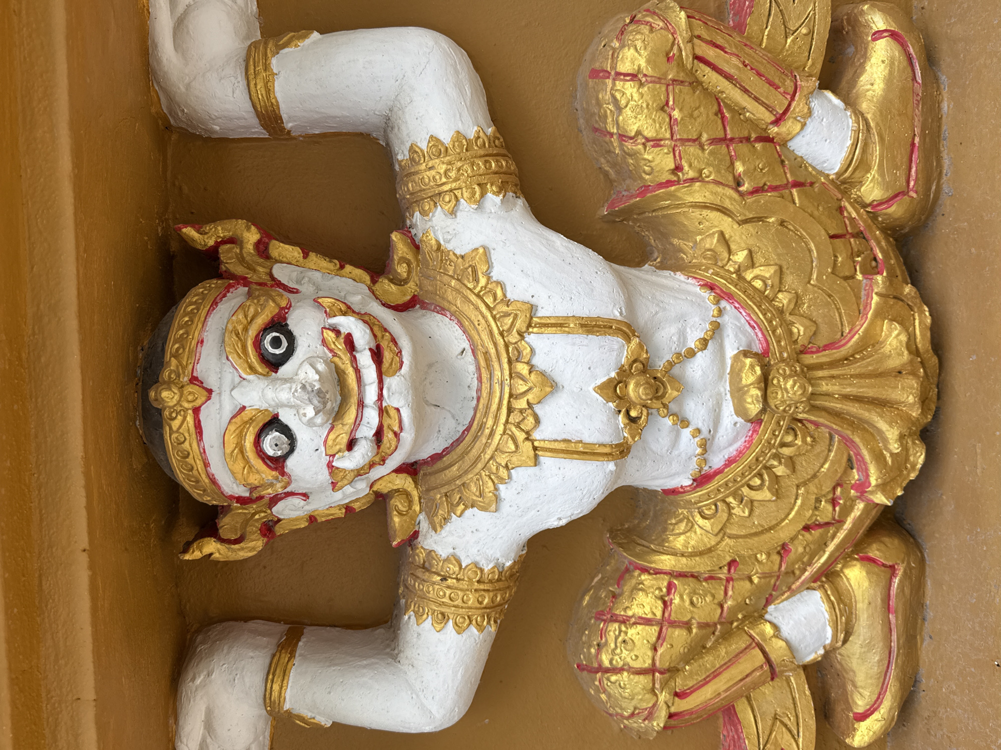 The image shows a close-up of a decorative statue of a mythical being, possibly a giant or demon, often found in temples or on traditional Thai architecture. The statue is painted white and adorned with intricate gold and red detailing. It has a fierce expression with wide eyes, a gaping mouth, and pointed ears. The figure is depicted in a crouching position, with its arms raised as if supporting a structure above. The gold embellishments include elaborate headdresses, necklaces, armbands, and ornate clothing around its legs and waist. The background is a plain, textured yellow wall, suggesting it's part of a larger building. This specific statue is located in Baan Tai, Thailand, a locale known for its blend of traditional and modern influences.
