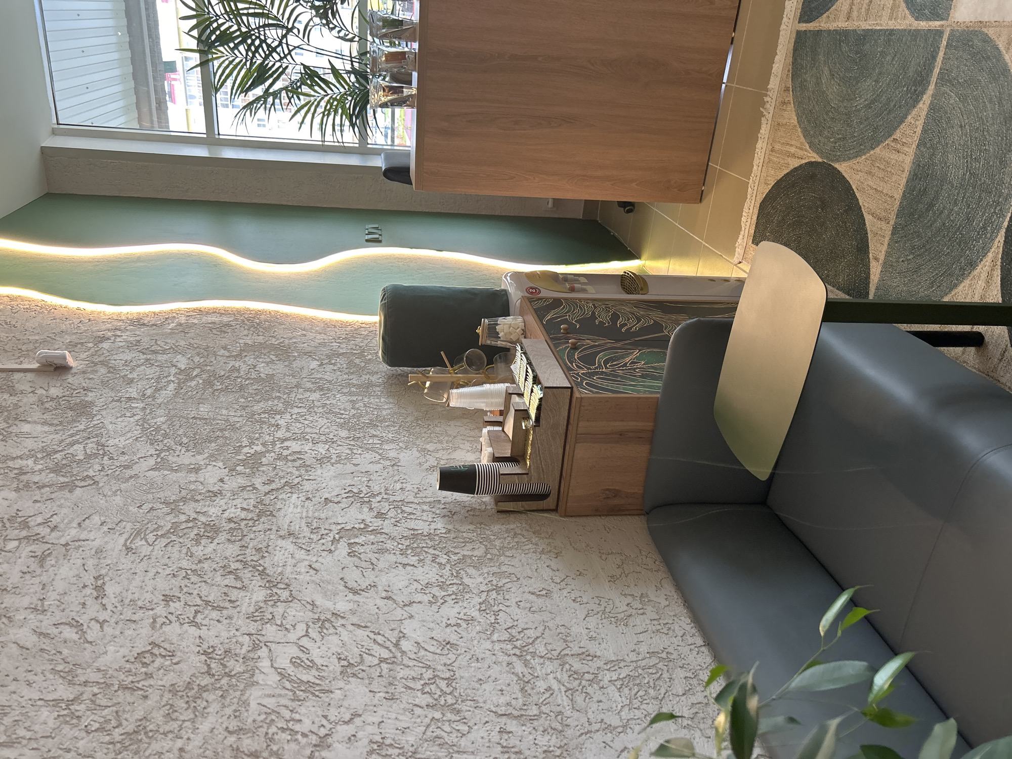 The image depicts an interior scene, likely a cafe or waiting area, in Medvedevo, Russia. The left side of the frame shows a textured, off-white wall with a decorative, wavy vertical light installation emitting a warm glow. In the foreground, a dark gray sofa with visible stitching detail is partially visible, with a small, metallic side table positioned on its right.
To the right of the sofa and wall, a wooden cabinet stands. On top of this cabinet is a wooden organizer holding stacks of paper cups and what appear to be sugar packets. A water cooler with a gold dispenser is positioned to the left of the cabinet. Behind the cabinet, a large, dark green object, possibly a cushion or upholstered item, is placed against the wall.
Further to the right, a wooden counter or desk is visible, with several clear glasses containing palm fronds. A large window is present on the far right, showing a view of buildings and foliage outside. The floor is tiled with light brown square tiles, and a rug with a geometric pattern of large, overlapping oval shapes in shades of beige and dark green is laid out in the foreground. The overall impression is of a modern, clean, and well-lit space.