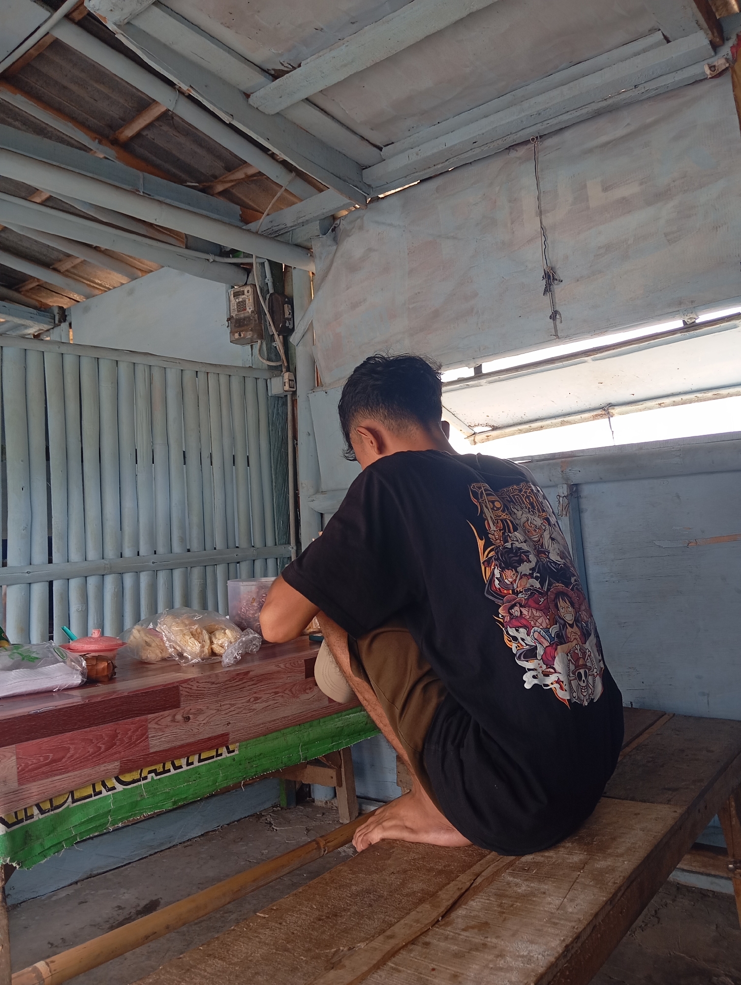 A young man wearing a black t-shirt with an anime graphic on the back is crouched on a wooden bench. He is facing away from the camera, towards a wooden table. On the table are several plastic bags of snacks and a small pink bowl. The scene appears to be outdoors, under a rustic wooden roof, possibly a stall or an open-air shelter. The background consists of light blue bamboo-like vertical panels. The overall environment suggests a humble, possibly rural setting in Sokaraja Wetan, Indonesia, perhaps near a market or a local eatery.