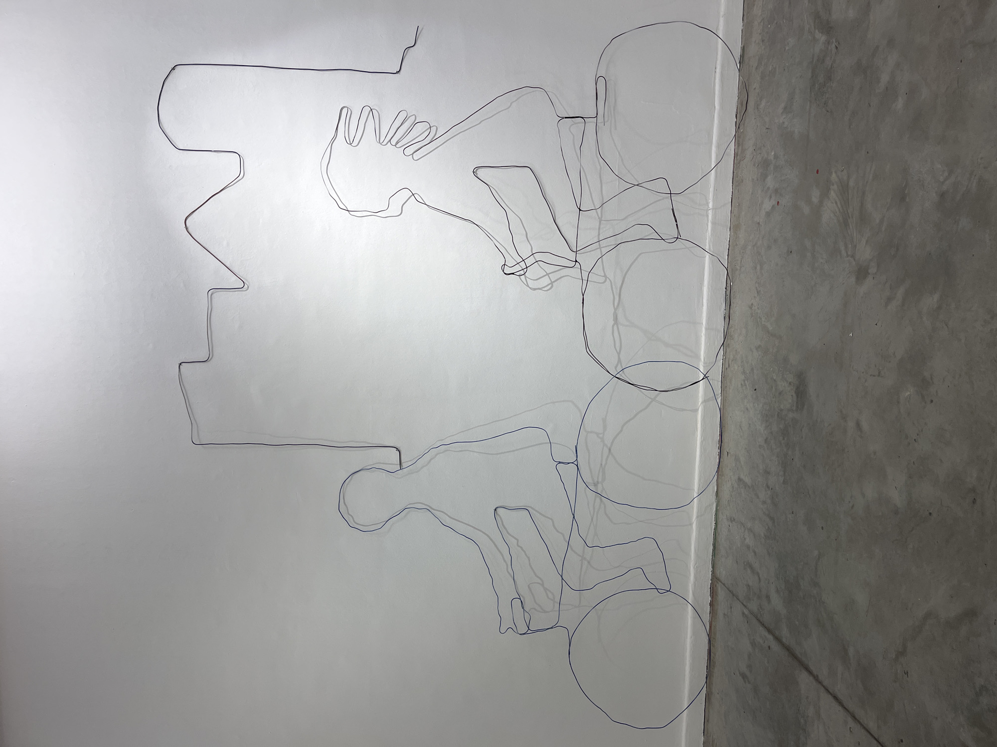 A wall with a wire art piece depicting a person riding a bicycle. The wire art has a shadow cast on the wall. The wall is white and adjacent to a concrete column. The location is likely indoors. It is not possible to determine the location is Buenos Aires, Argentina from the image.