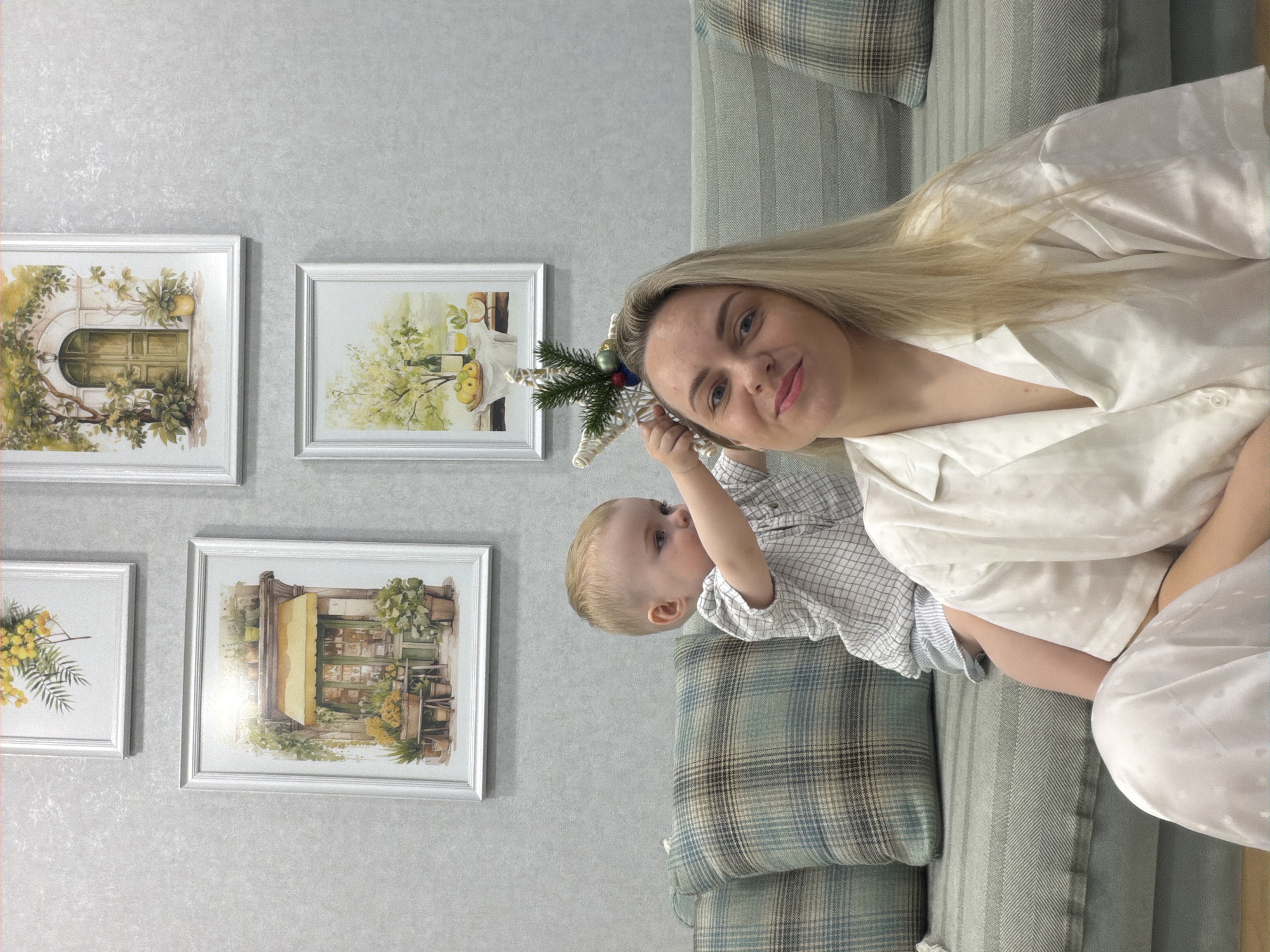 An indoor, vertically oriented portrait captured in Votkinsk, Russia, shows a blonde woman and a baby seated on a sofa. The woman, with fair skin and light blue eyes, wears a white, satiny shirt or pajamas and smiles gently towards the viewer. She holds a baby, likely a toddler, who is wearing a light-colored checkered shirt. The baby is engrossed in playing with a white, star-shaped Christmas tree topper adorned with small pine branches and miniature ornaments.
They are positioned on a sofa featuring plaid and striped cushions in shades of blue, green, and beige. Behind them, a light gray wall with a subtle textured pattern is decorated with four framed pictures arranged in a grid-like fashion. These watercolor-style artworks depict idyllic scenes: an arched doorway with greenery, a still life with fruit and a bottle, a charming storefront with an awning and potted plants, and a branch of yellow mimosa flowers. The lighting suggests daytime. The overall mood of the image is tender and domestic, highlighting a moment of familial interaction. No visible text is present in the image.