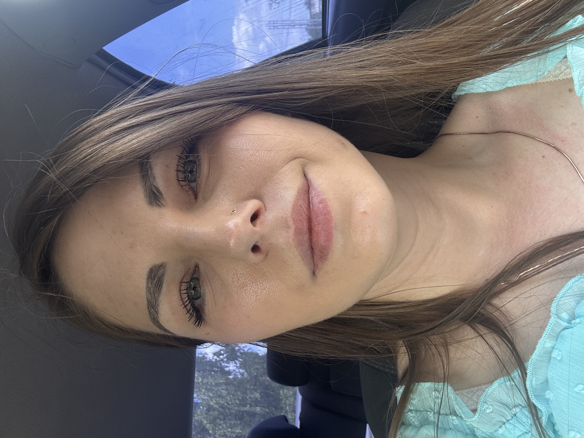 A woman with light brown hair and green eyes is posing for a selfie in a car. She has a nose piercing and is wearing a light blue top. The car has a sunroof, and the sky is visible through it. The image does not contain sufficient information to determine a location in Shevlyakovo, Russia.