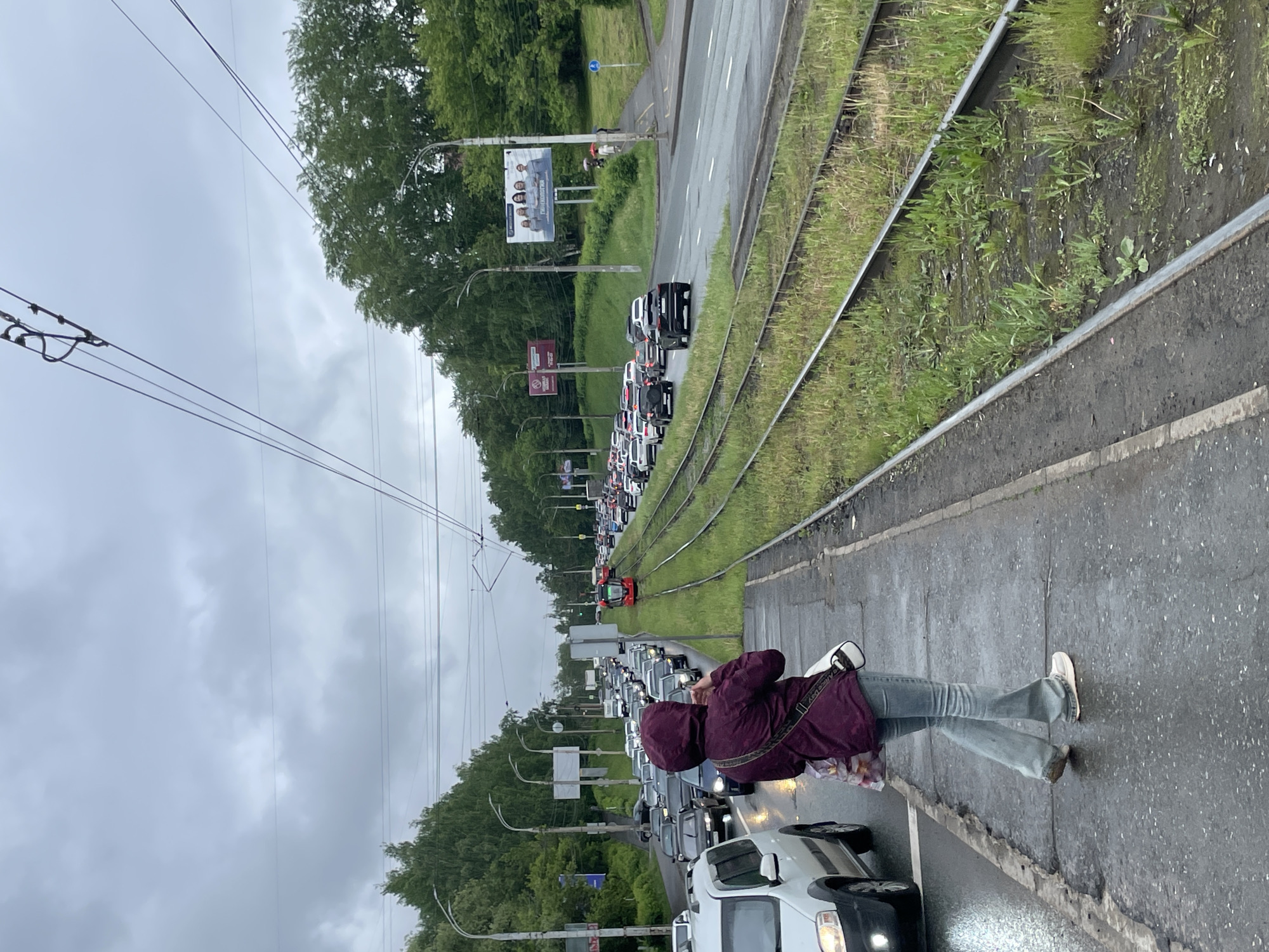 The image shows a traffic jam in Izhevsk, Russia on a road lined with tram tracks. A person in a hooded purple jacket, jeans, and white shoes is standing on the sidewalk near the road. They are looking at their phone. The tram tracks are overgrown with grass and weeds. Overhead tram lines are visible, as well as several streetlights. The sky is overcast. A billboard is visible in the distance along with numerous cars stuck in traffic.