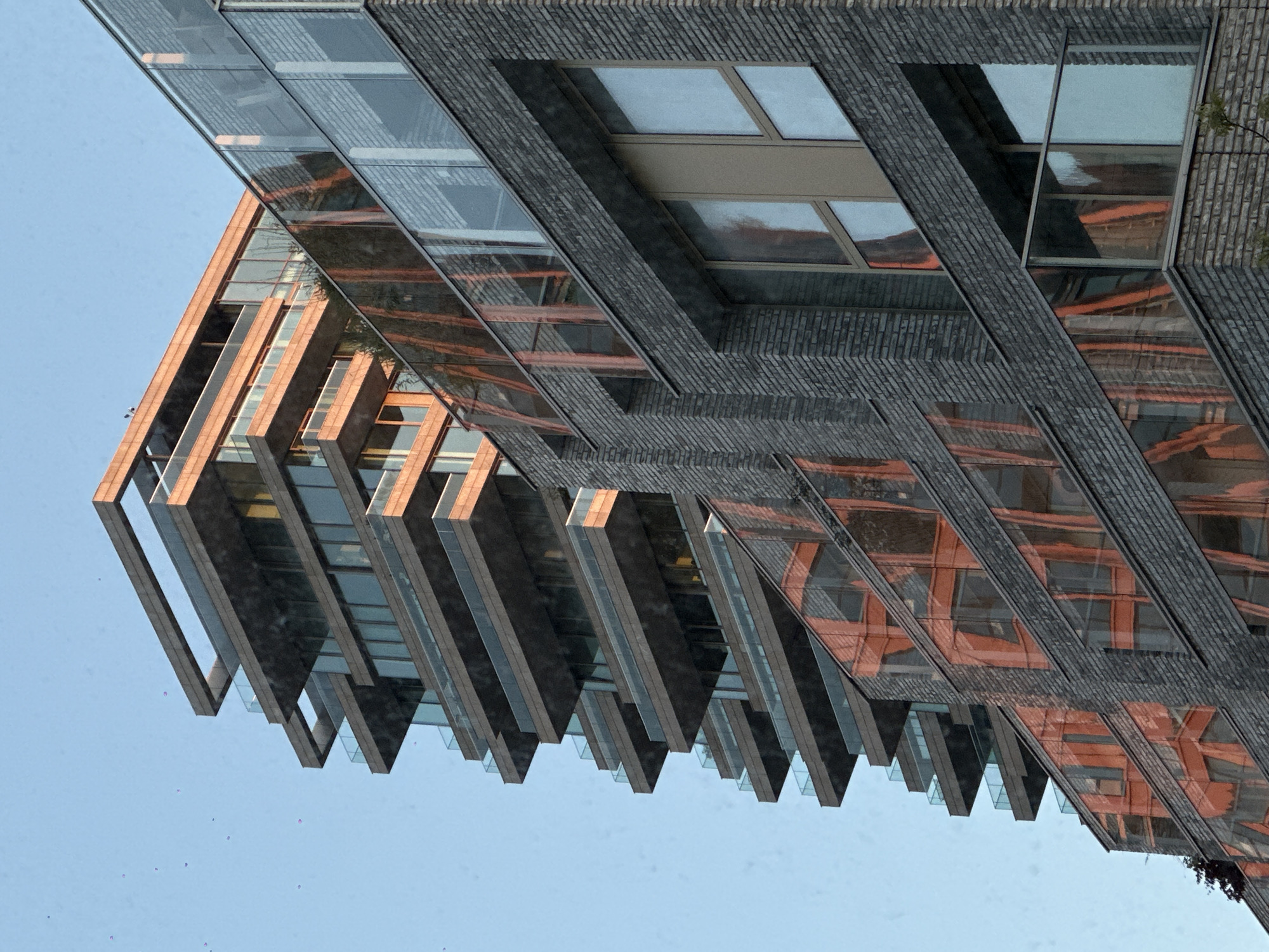 A modern, multi-story building with a distinctive architectural design is captured against a clear, light blue sky. The building primarily features sections of dark grey brickwork, laid in horizontal patterns, interspersed with extensive reflective glass facades.
The structure is characterized by a series of angular, cantilevered elements that protrude outwards, particularly on the left side of the image. These sections have dark grey or black frames and undersides in a contrasting warm, reddish-brown or copper-like material, incorporating glass panels that reflect the surrounding environment and parts of the building itself. The reflections, especially those of the reddish-brown elements, create complex, fragmented geometric patterns across the glass surfaces.
The sky is uniformly clear and cloudless, indicating daytime with good weather. Small, faint purple specks are sparsely scattered against the blue sky in the lower-left area. A small amount of dark green foliage is visible on the far right edge, suggesting nearby vegetation. No people or specific activities are depicted within the frame. There is no visible text. While the location is provided as Amsterdam, Netherlands, the image itself does not contain explicit geographical markers.