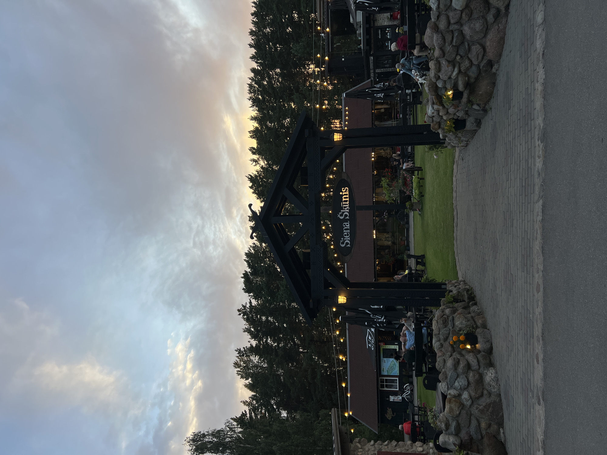 The image depicts an outdoor seating area at a restaurant or establishment named "Siena Skūnis" in Pērkone, Latvia. The setting appears to be at dusk, with a soft, overcast sky displaying hues of blue, gray, and hints of orange. A line of tall pine trees forms a natural backdrop to the scene.

In the foreground, a paved walkway leads towards a rustic, dark wooden archway adorned with string lights. Flanking the walkway are low, stone-built walls containing small, decorative planters with orange flowers and small lights.

Under the archway, the establishment's name, "Siena Skūnis," is visible on a sign. Beyond the archway, several people are seated at tables, some under umbrellas, enjoying the evening. The area is furnished with dark tables and chairs, and umbrellas are open over some of the seating areas, suggesting a place of dining or relaxation. A few buildings with dark, sloping roofs are visible behind the outdoor seating area. The overall atmosphere is calm and relaxed, with the evening light casting a warm glow.
