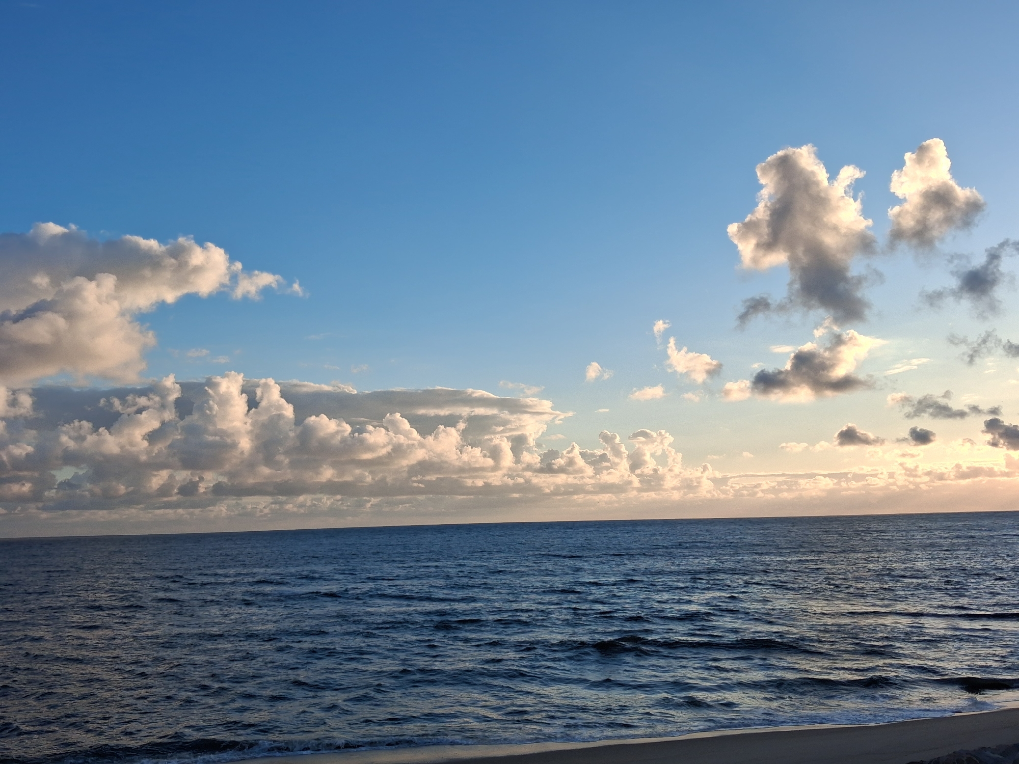 The image shows a coastal scene at Figueira da Foz, Portugal. A body of water, likely the Atlantic Ocean, fills the foreground. Small waves are visible. A narrow strip of beach is present at the bottom of the frame. The sky takes up the upper half of the image. There are clouds in the sky, varying in size and density, with sunlight filtering through some of them. The overall lighting suggests either sunrise or sunset.